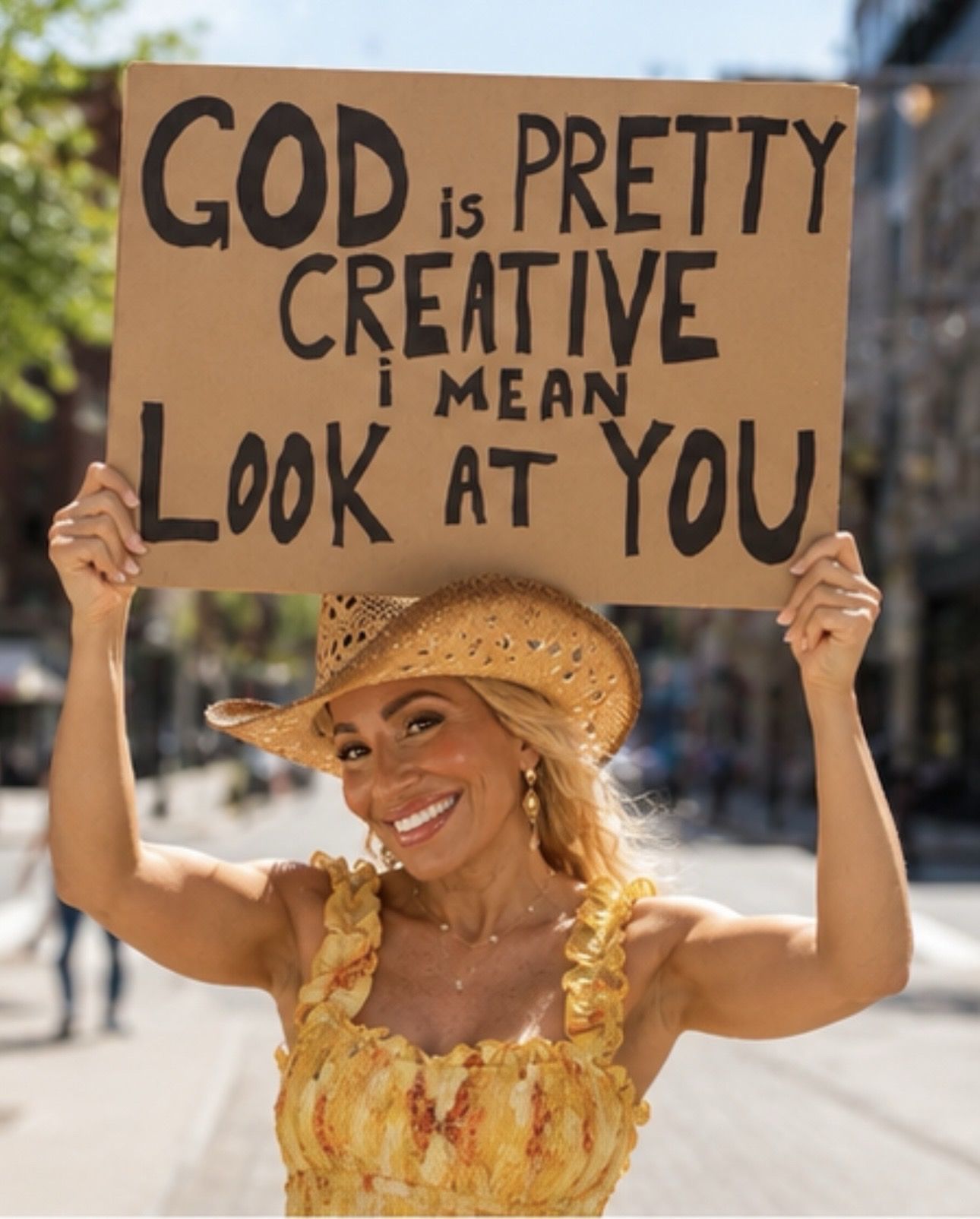 Woman smiling, wearing a straw hat and yellow dress, holding a sign that says God is pretty, creative, and looks at you.