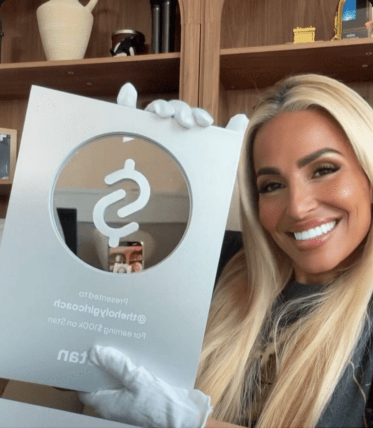 Woman with long blonde hair smiling, holding a framed mirror with a medical symbol, in a room with wooden shelves.