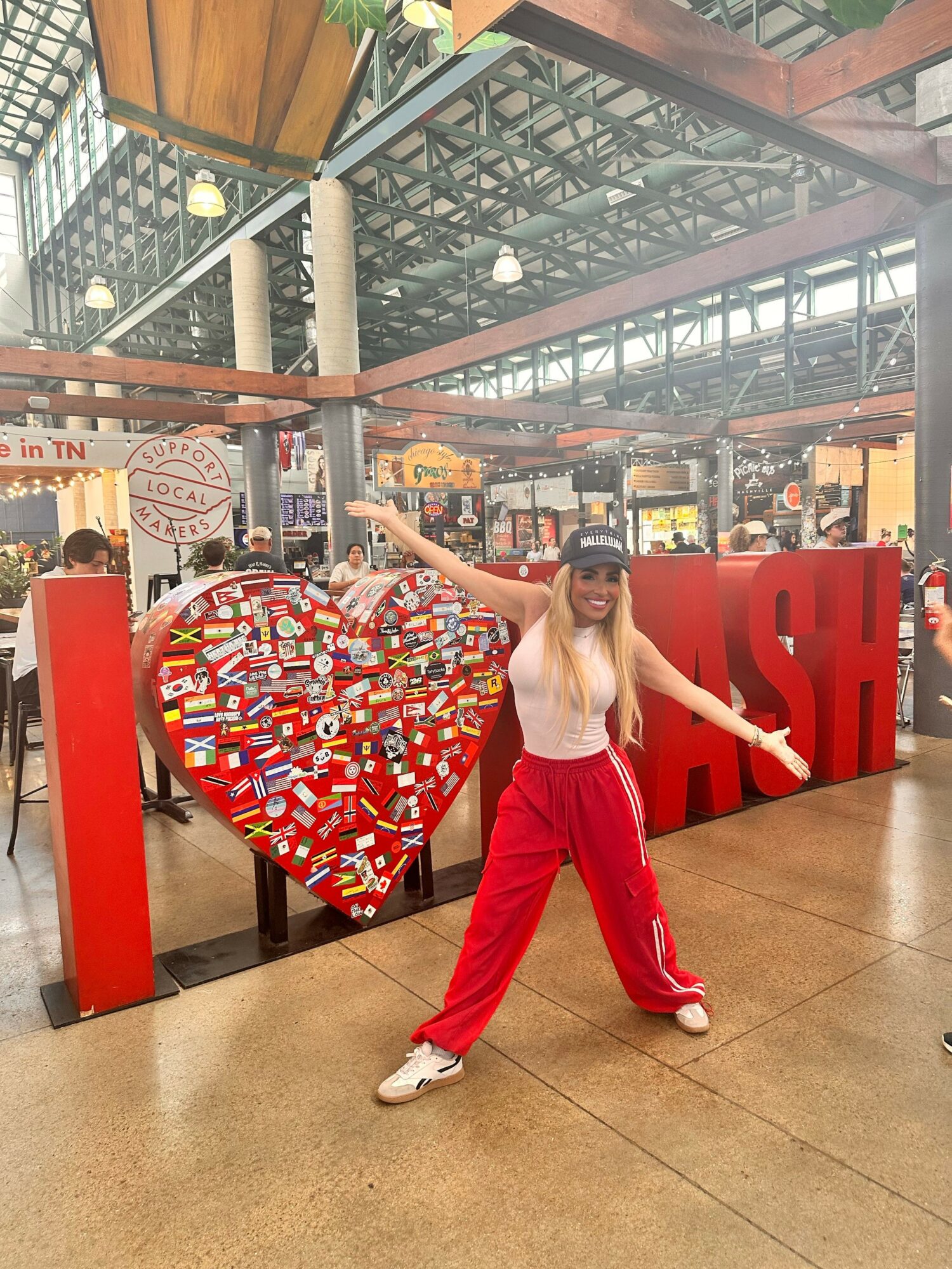 Person with long hair, wearing a cap, white top, and red pants, standing in front of a large heart and letters in a shopping mall.