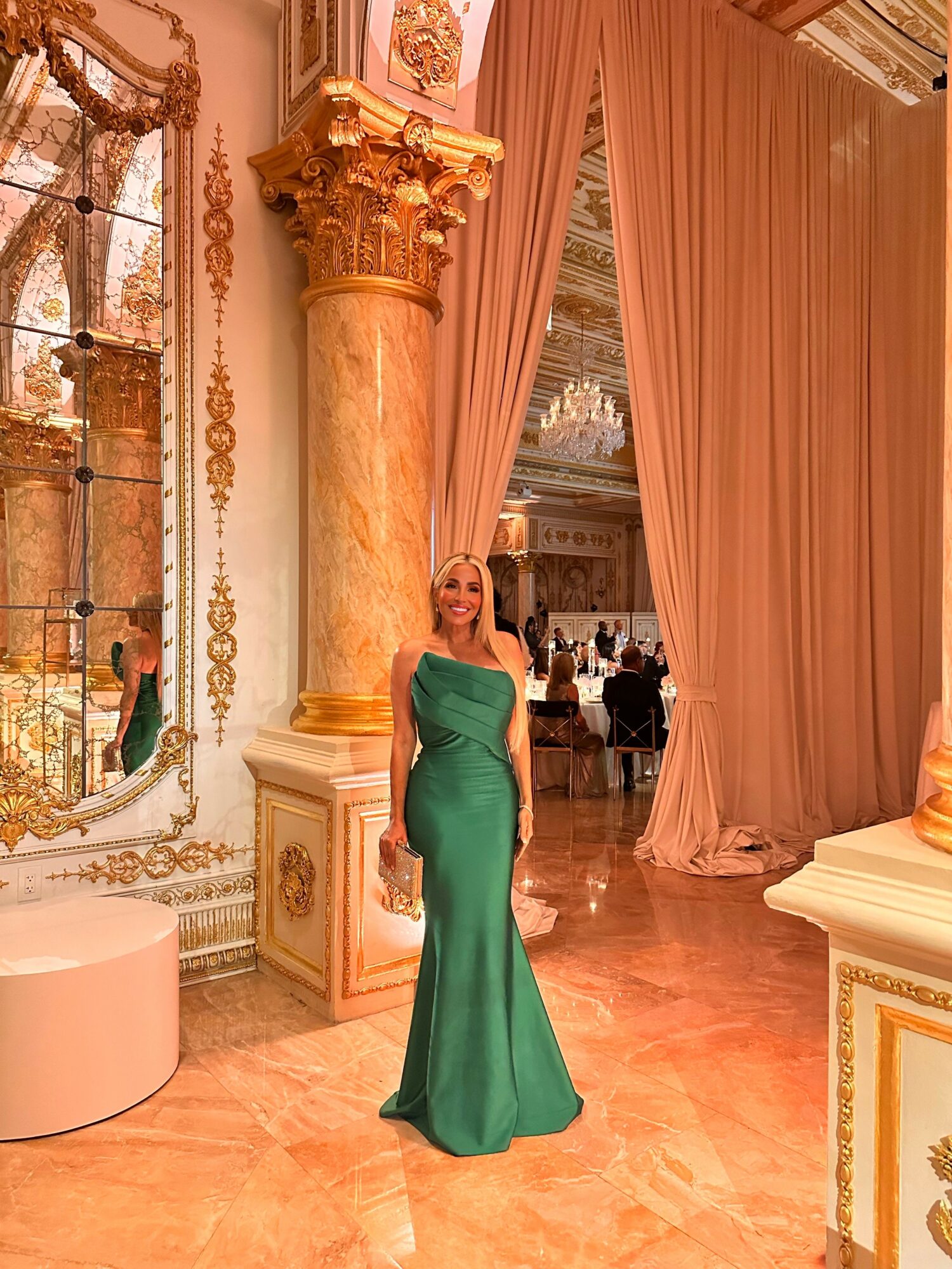 Woman in a green gown standing in a luxurious, ornate room with gold accents and draped curtains.