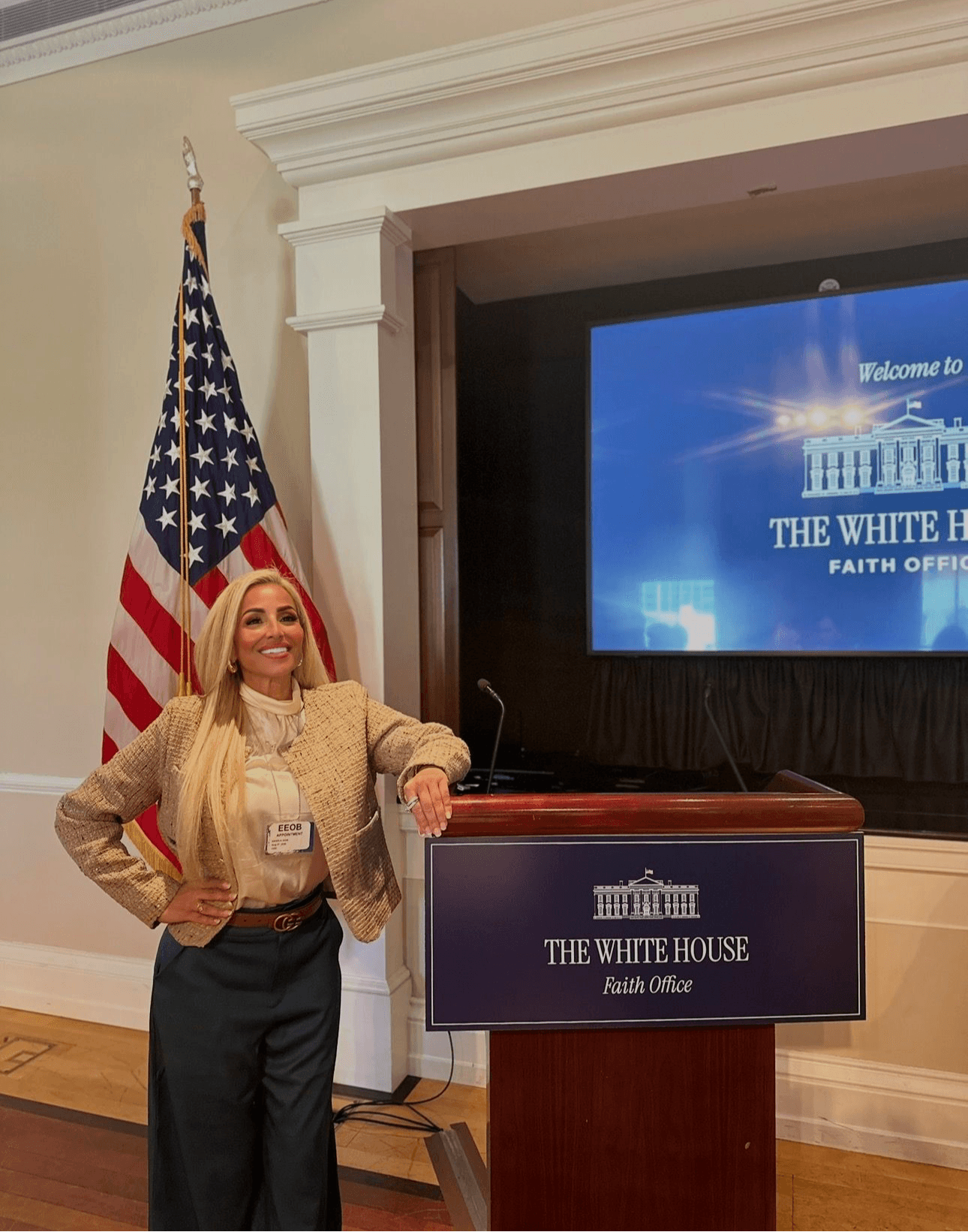 Woman standing next to a podium and American flag in a room with a large screen displaying the White House logo.