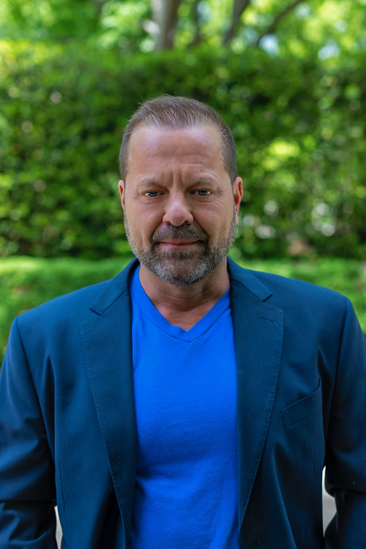 Man with short hair and beard wearing a blue shirt and blazer outdoors with green trees in background.
