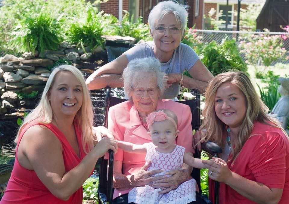Four women and a young girl outdoors, smiling, with greenery and a stone wall in the background.