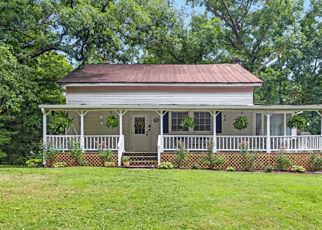 White house with a porch, surrounded by trees and a grassy yard, with hanging plants on the porch.
