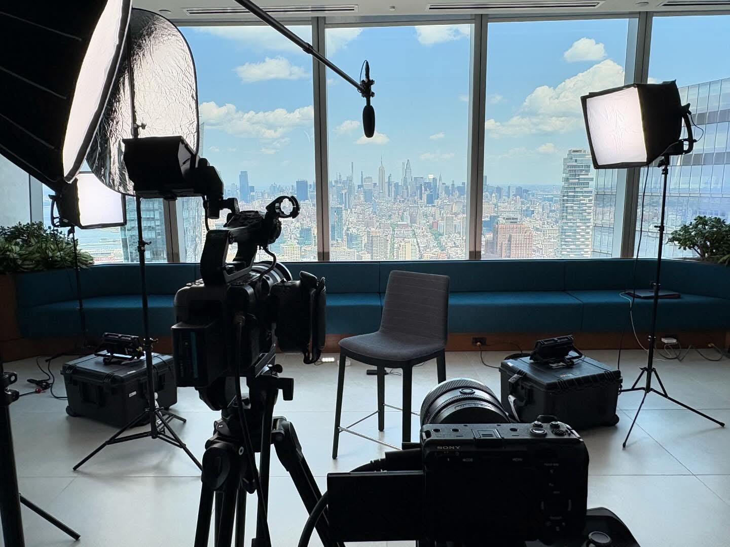 Studio setup with camera, lights, and a chair in front of large windows showing a cityscape.
