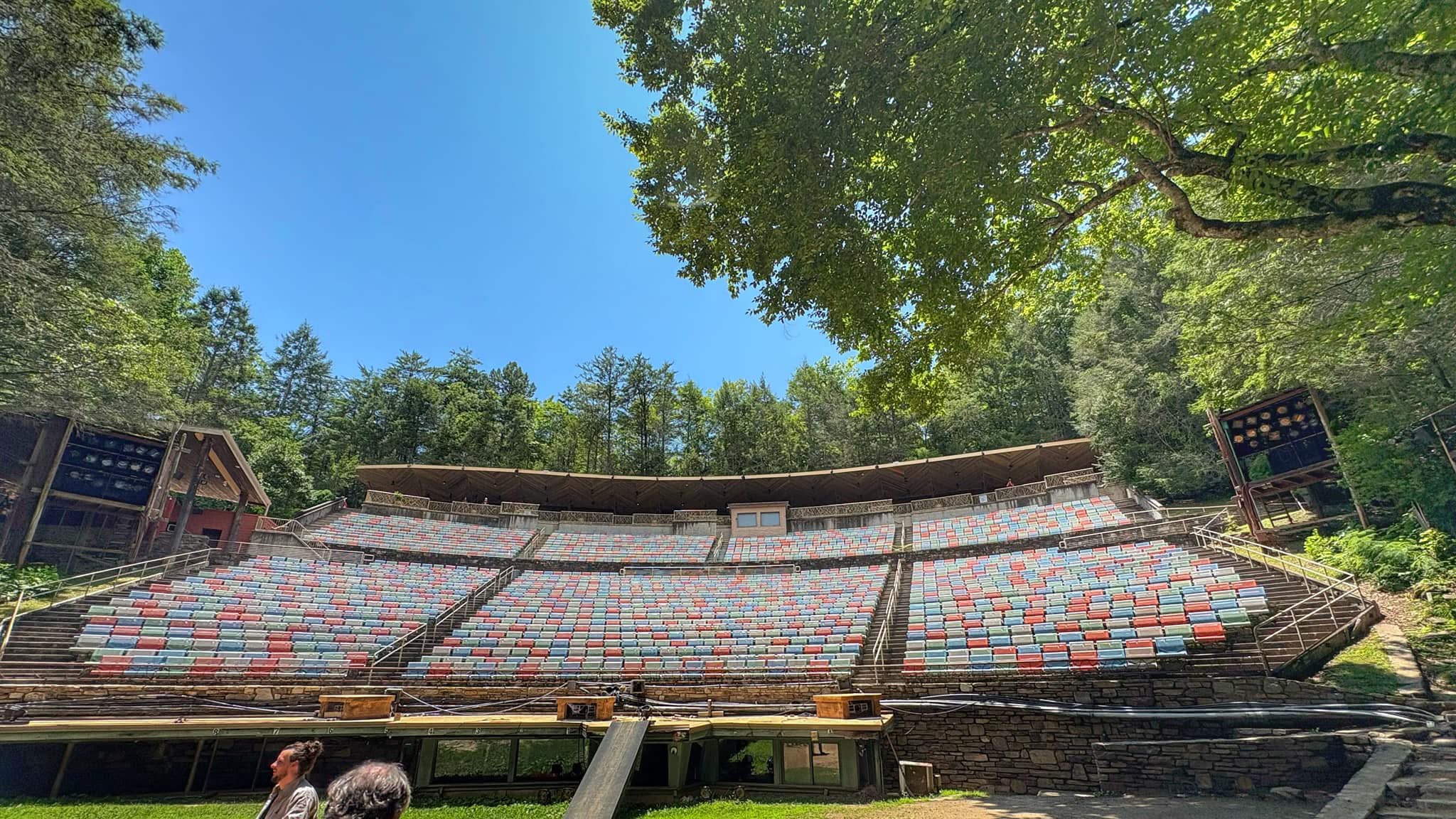 Empty outdoor theater with rows of seats, surrounded by trees and a clear blue sky.
