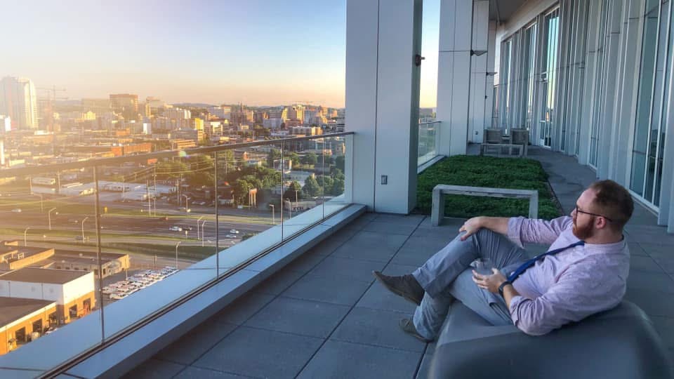Man sitting on a balcony overlooking a cityscape during sunset, using a laptop, with glass walls and modern furniture.