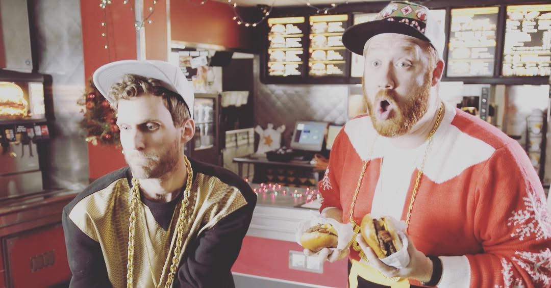Two men in a fast-food restaurant, one wearing a hat and gold jacket, the other in a red jacket and gloves, holding food.