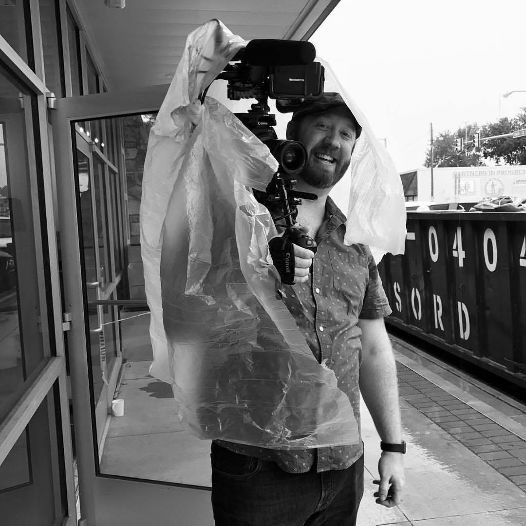 Person holding camera with raincoat hood, smiling, standing on train platform, black and white photo.