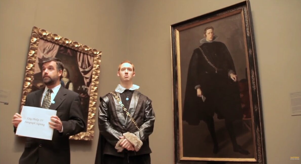 Two people stand in front of paintings in a gallery, one holding a sign, the other with hands clasped, a large portrait behind them.