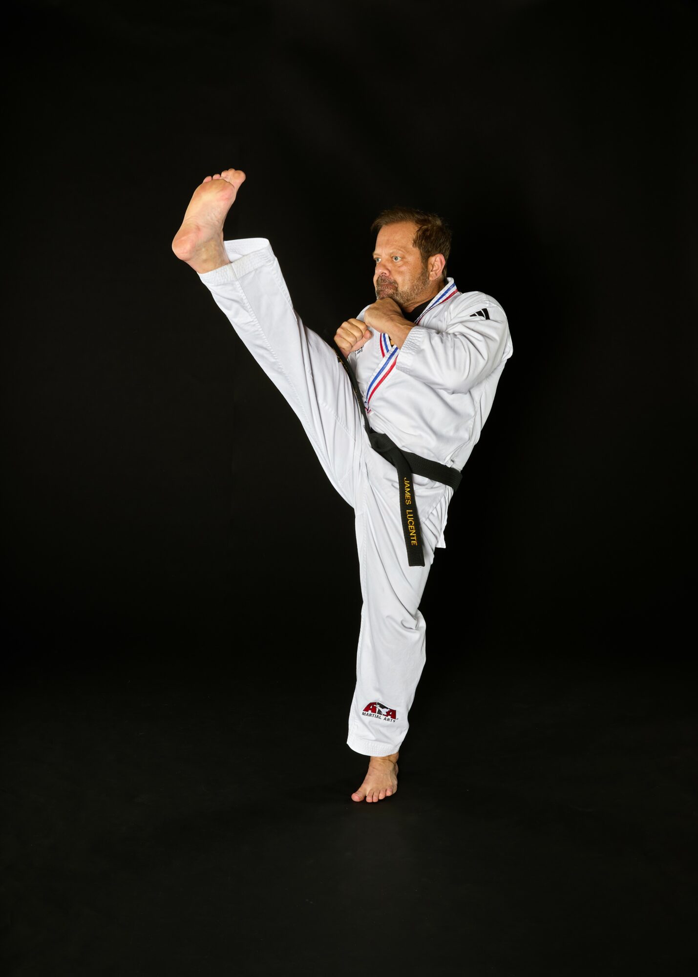 Martial artist performing a high kick against a black background, wearing a white uniform with a black belt.
