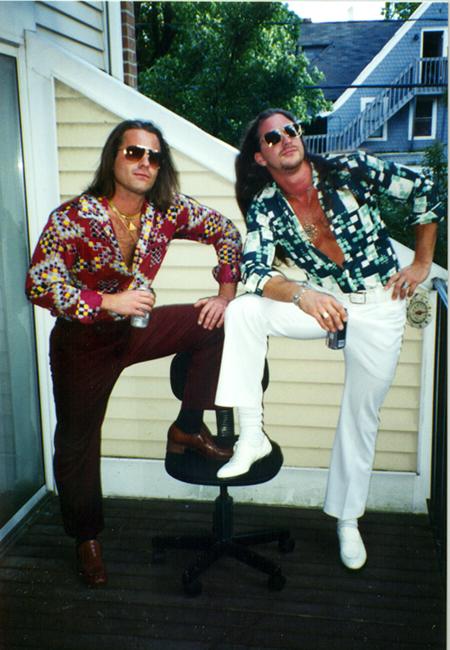 Two men with long hair and sunglasses pose outdoors, one with a patterned shirt and the other with a floral shirt, against a house wall.