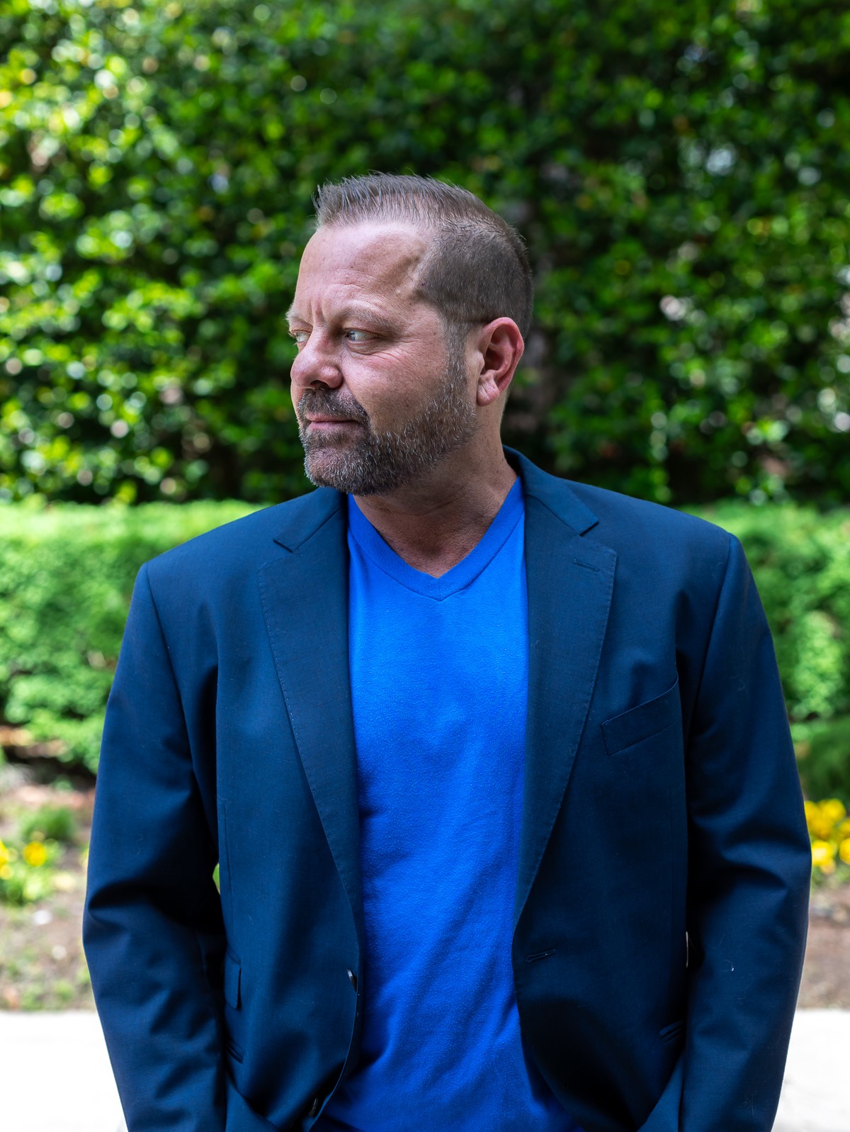 Man with short hair and beard wearing a blue shirt and blazer outdoors, with green foliage background.