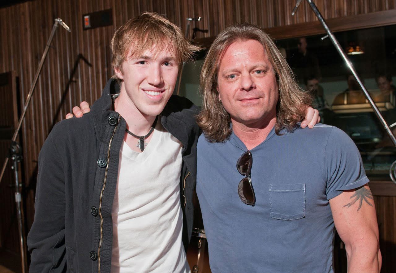 Two men with shoulder-length hair and casual clothing pose together indoors, smiling at the camera.