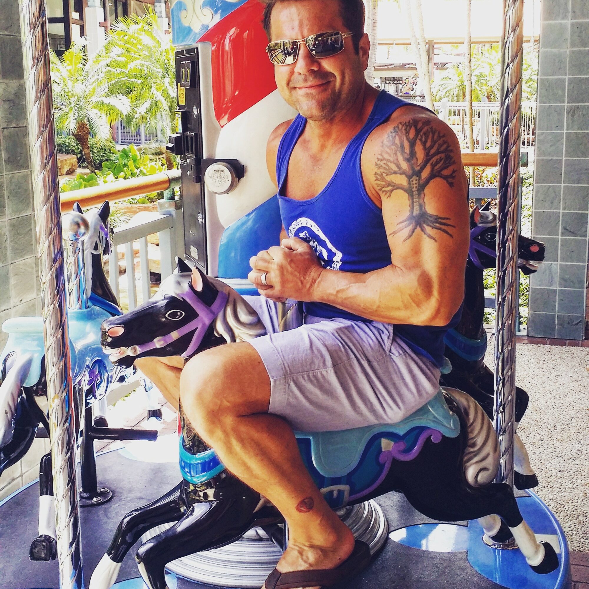 Man sitting on a mechanical horse ride, smiling, wearing sunglasses, a blue tank top, and shorts, with tattoos on his arm.