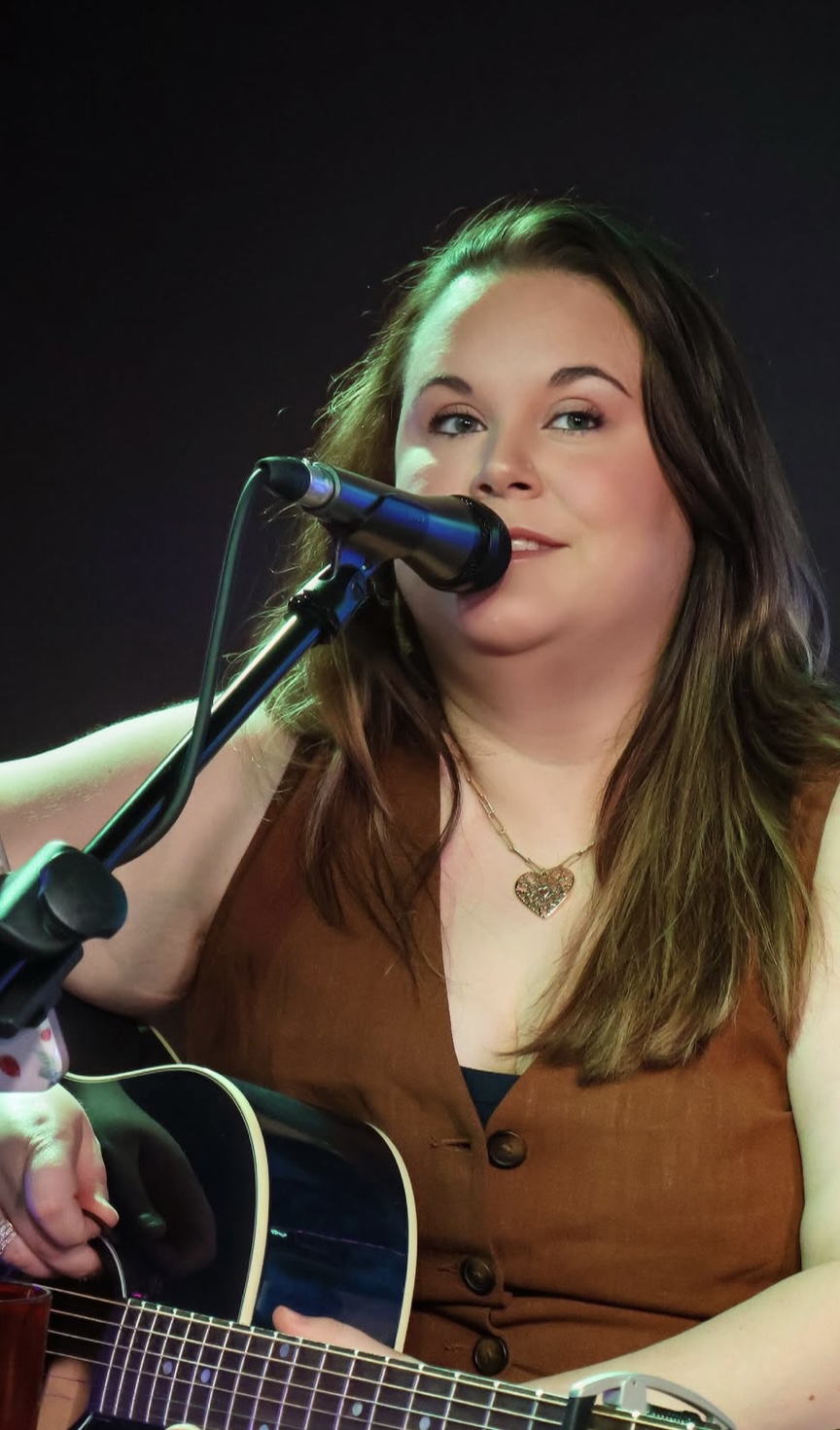 Woman singing into microphone while playing guitar, wearing a brown top and necklace, with dark background.