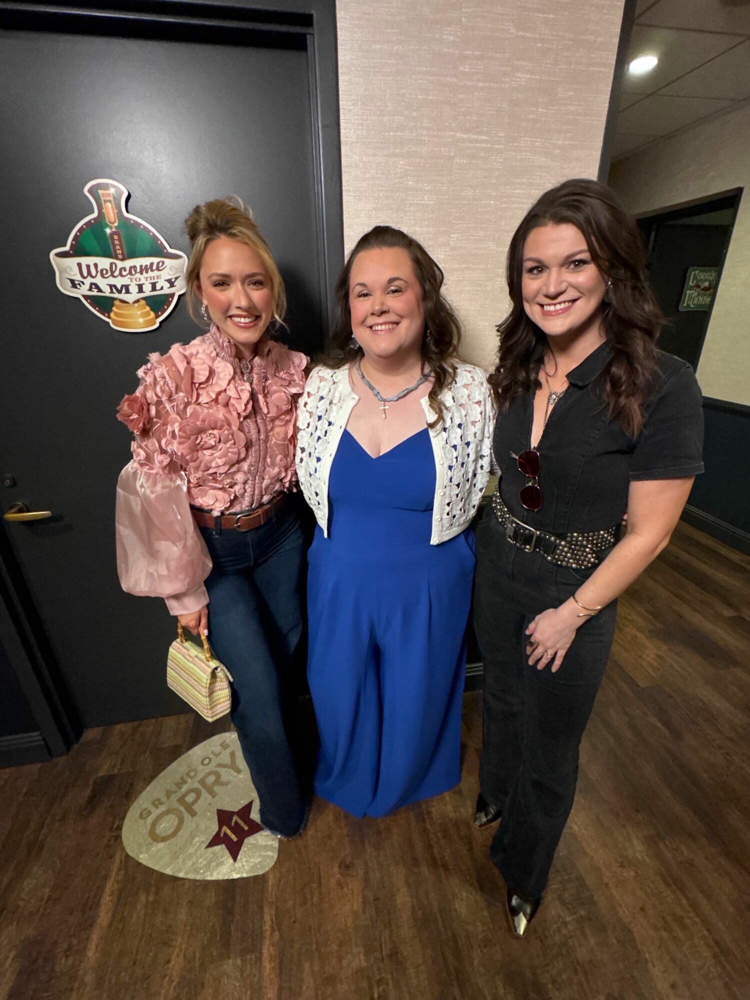 Three women standing indoors, one holding a small gift, with a logo on the floor and a dark wall behind.