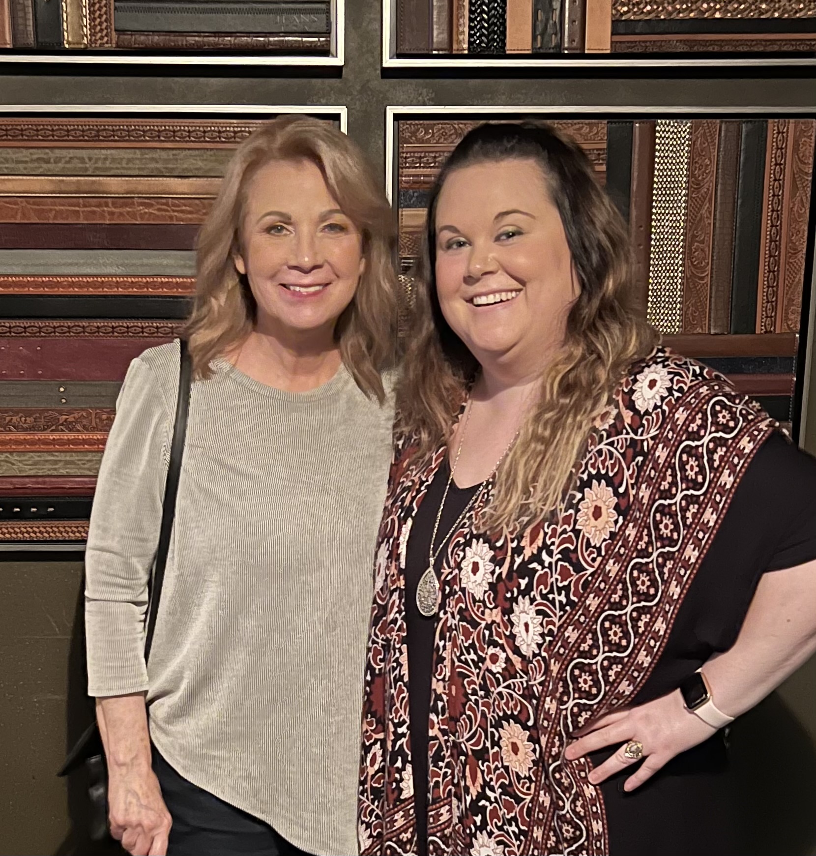 Two women smiling, standing in front of a bookshelf filled with books, one wearing a light gray top, the other a patterned shawl.