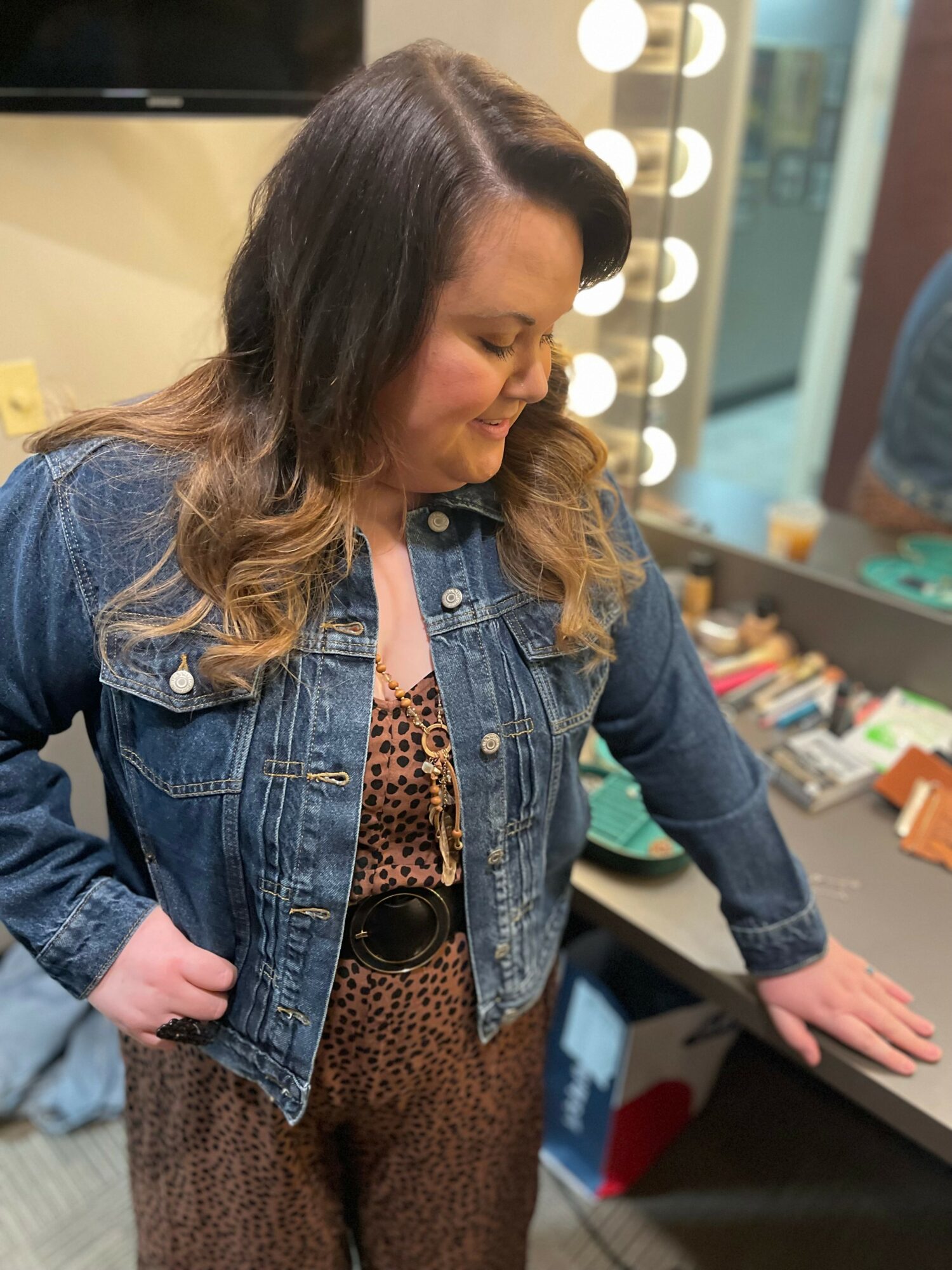Woman with wavy hair in a denim jacket and leopard print outfit, standing near a mirror with lights, in a room with books and objects.