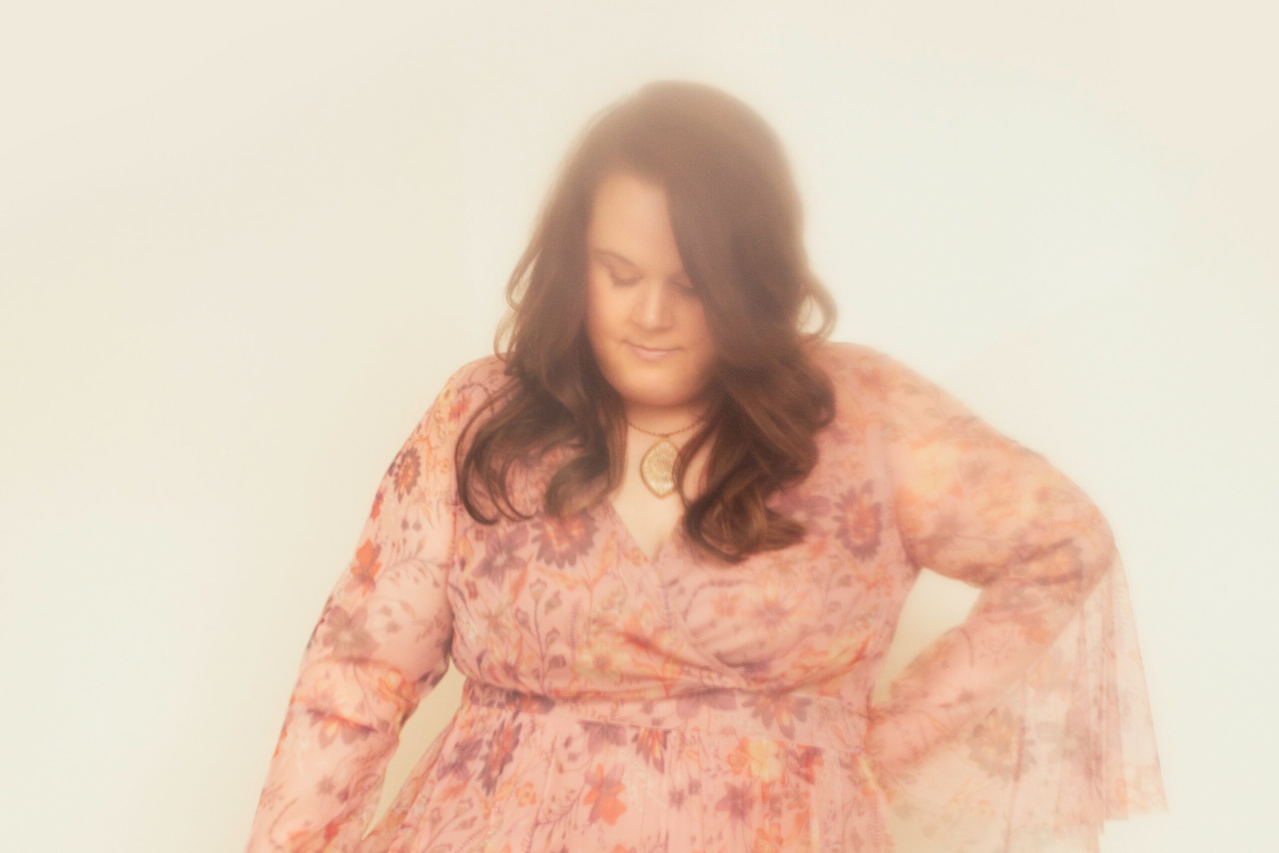 Woman with long dark hair wearing a pink floral dress, standing with one hand on her hip, smiling softly.