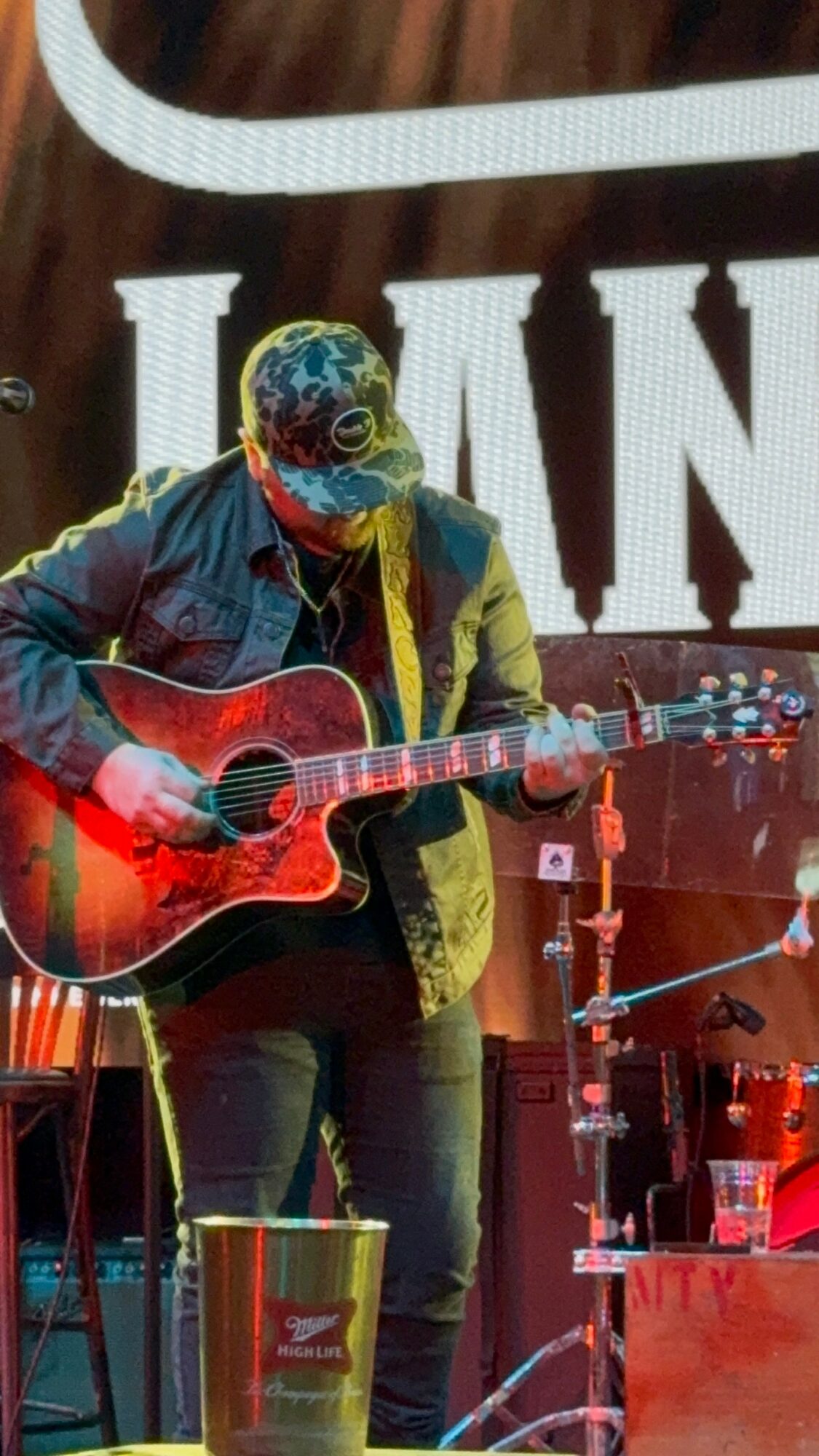 Person playing guitar on stage with large letters in background, wearing a cap and jacket, illuminated by stage lights.