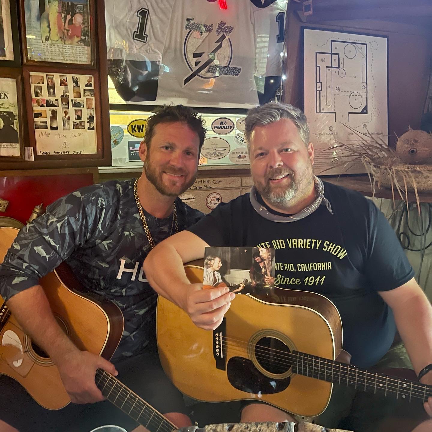 Two men sitting with guitars, smiling, in a room with framed pictures and posters on the wall.