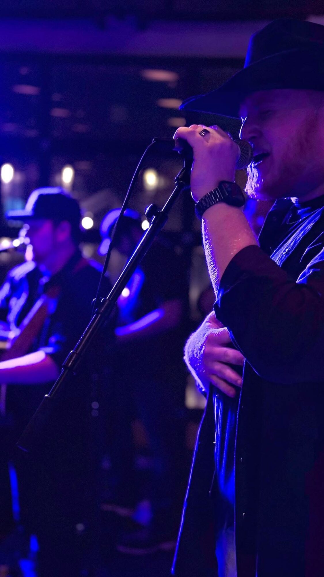 Person singing into a microphone on stage with a band in the background, dimly lit with blue and purple lights.