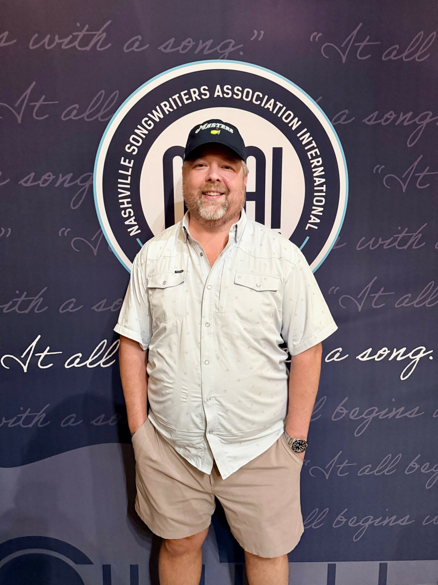 Smiling man in casual clothing standing in front of Nashville Songwriters Association International backdrop.