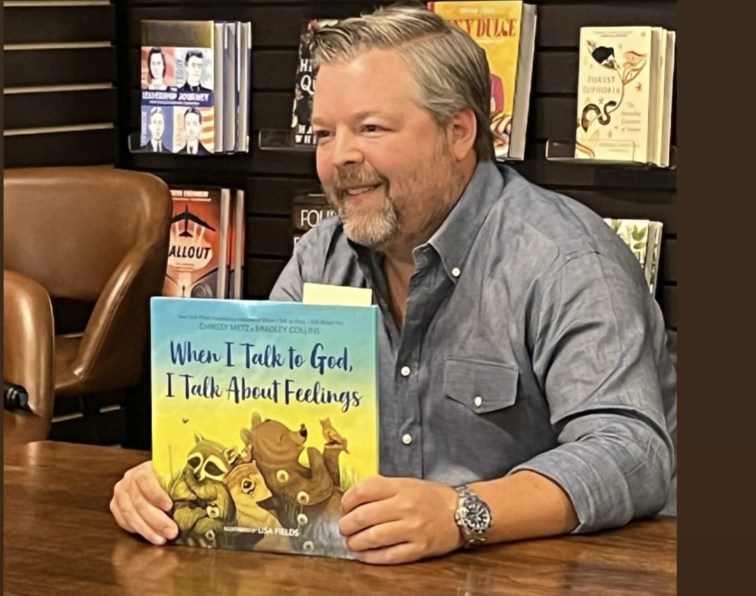 Man with gray hair and beard sitting at a table holding a children's book titled 'When I Talk to God, I Talk About Feelings' in a bookstore.