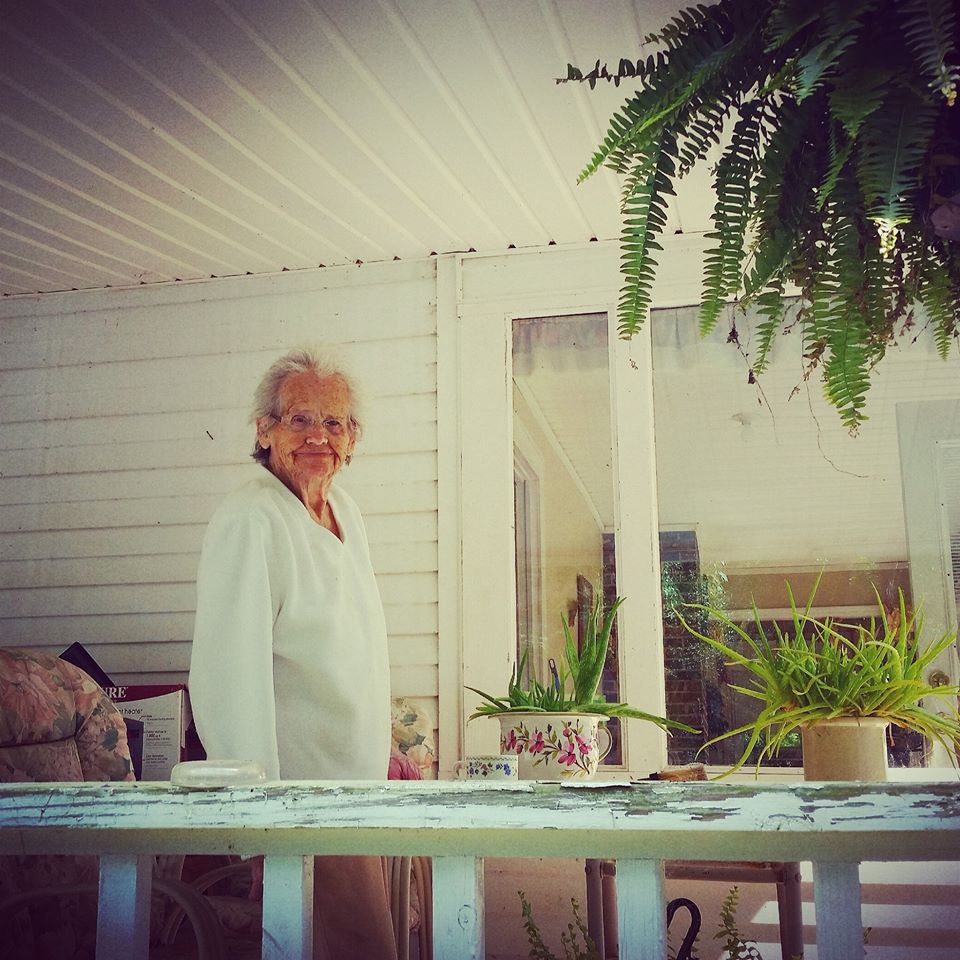 Elderly woman with white hair and glasses standing indoors near a window, surrounded by potted plants and a table.