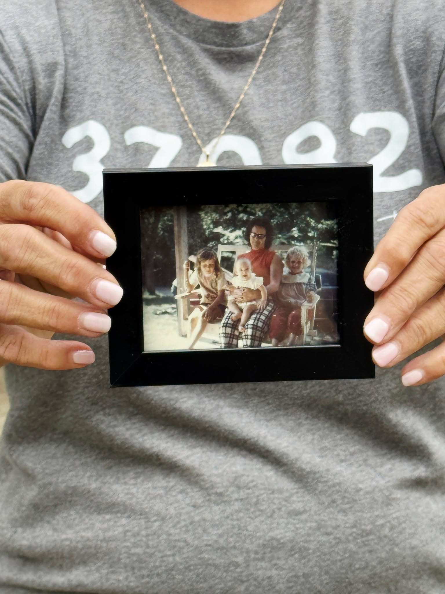 Person holding a framed photo of children sitting outdoors on a bench, with trees in the background.