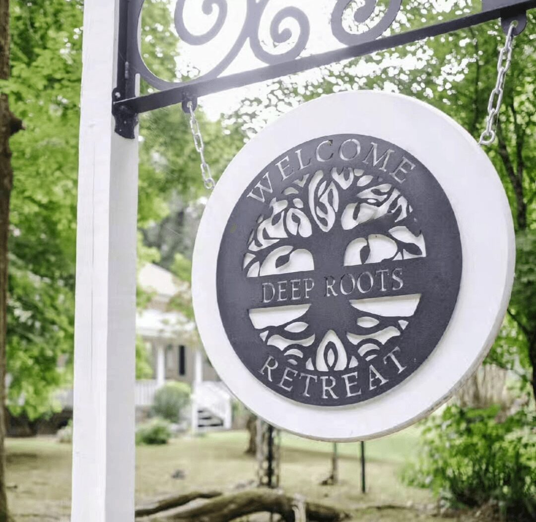 Round sign hanging outdoors with a tree design and text, reading 'Welcome Deep Roots Retreat,' against a background of trees and houses.