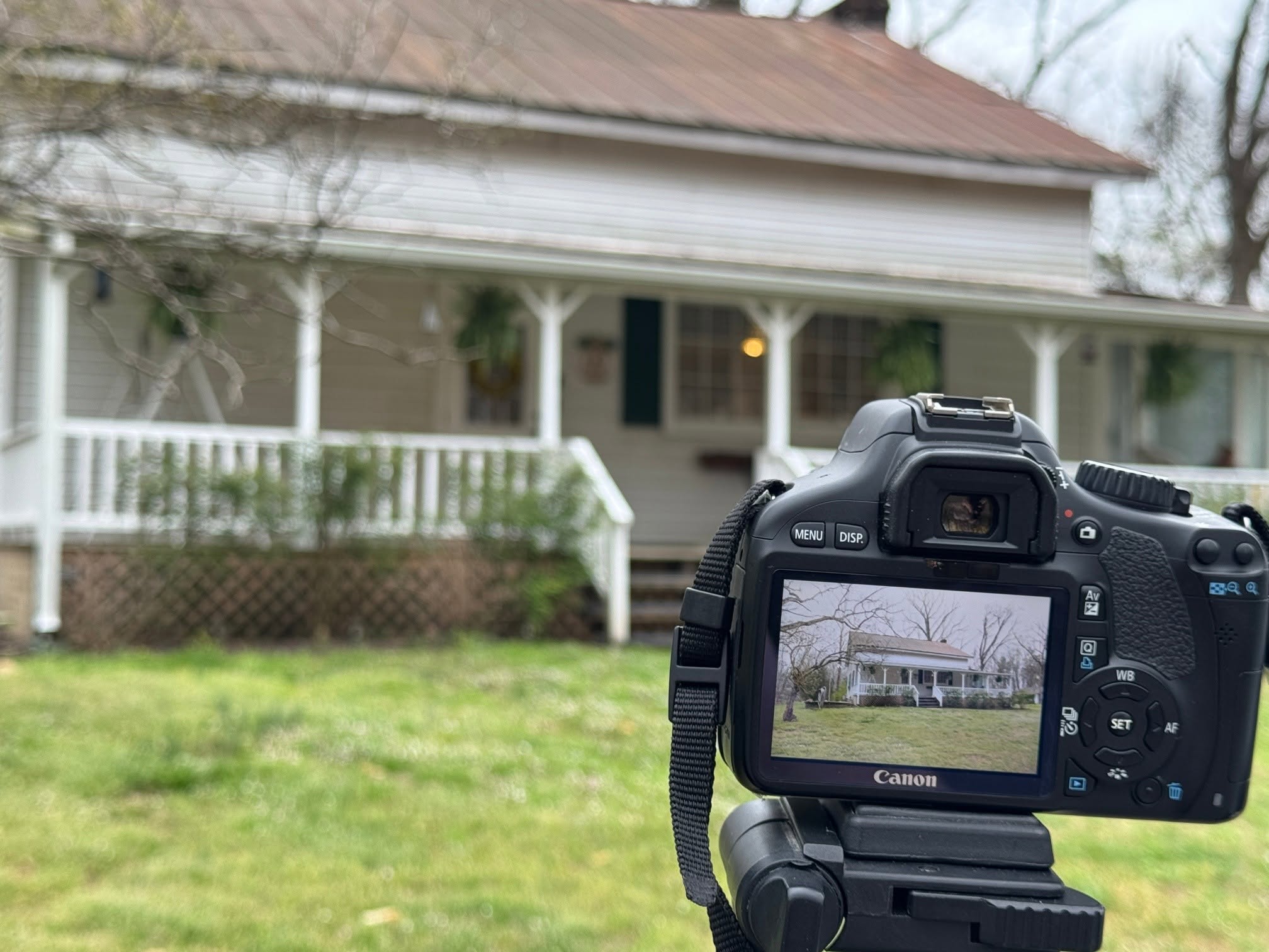Camera view showing a house with a porch and white railing, surrounded by grass and trees, under an overcast sky.
