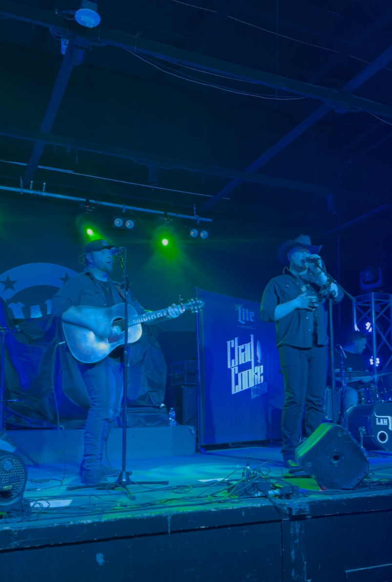 Two musicians perform on stage with green and blue lighting, one playing guitar and the other singing into a microphone.