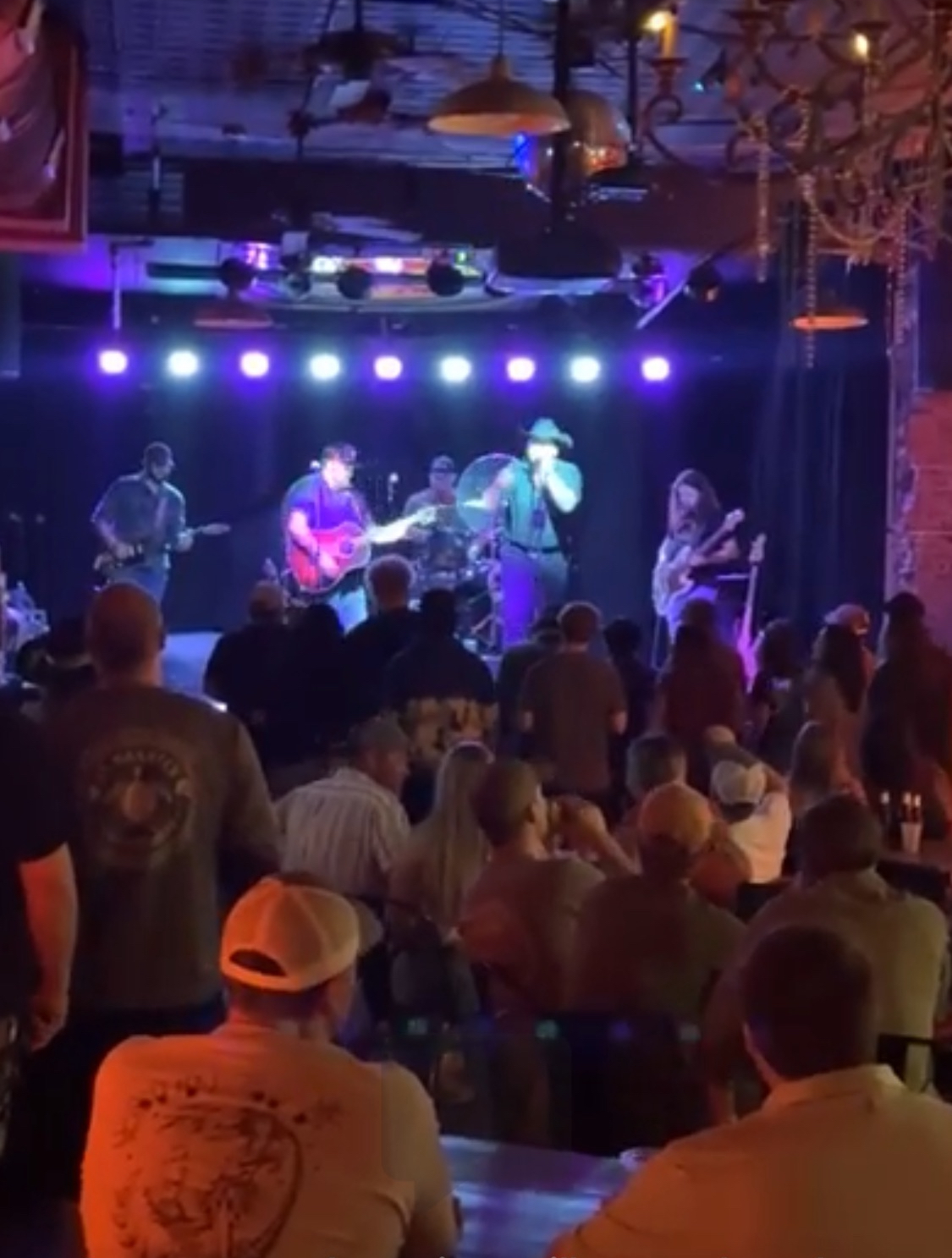 Band performs on stage with audience watching, colorful stage lights illuminate the scene, indoor venue with ceiling decorations.