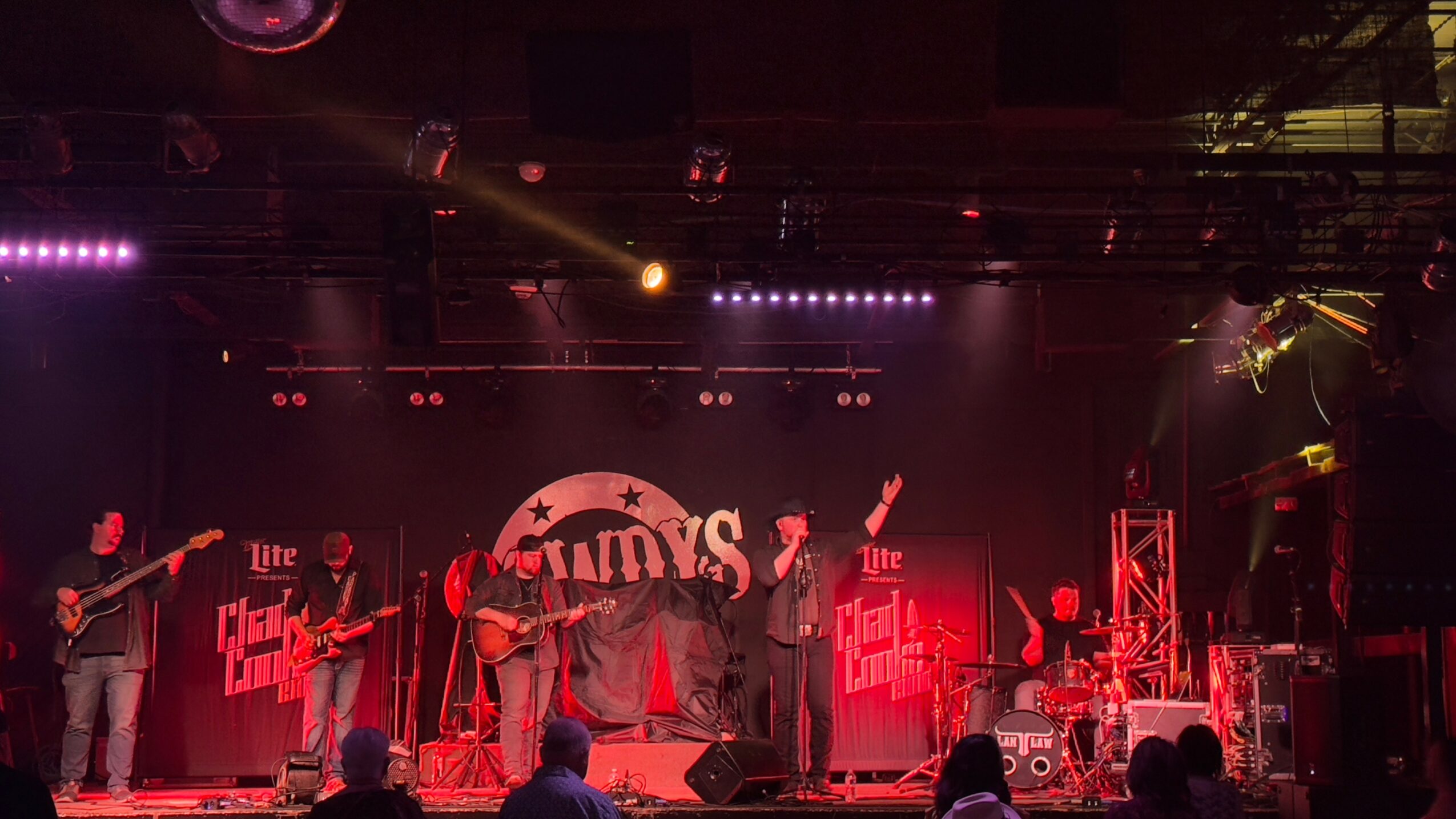 Band performing on stage with red and purple lighting, audience visible in foreground, large backdrop with text and graphics.