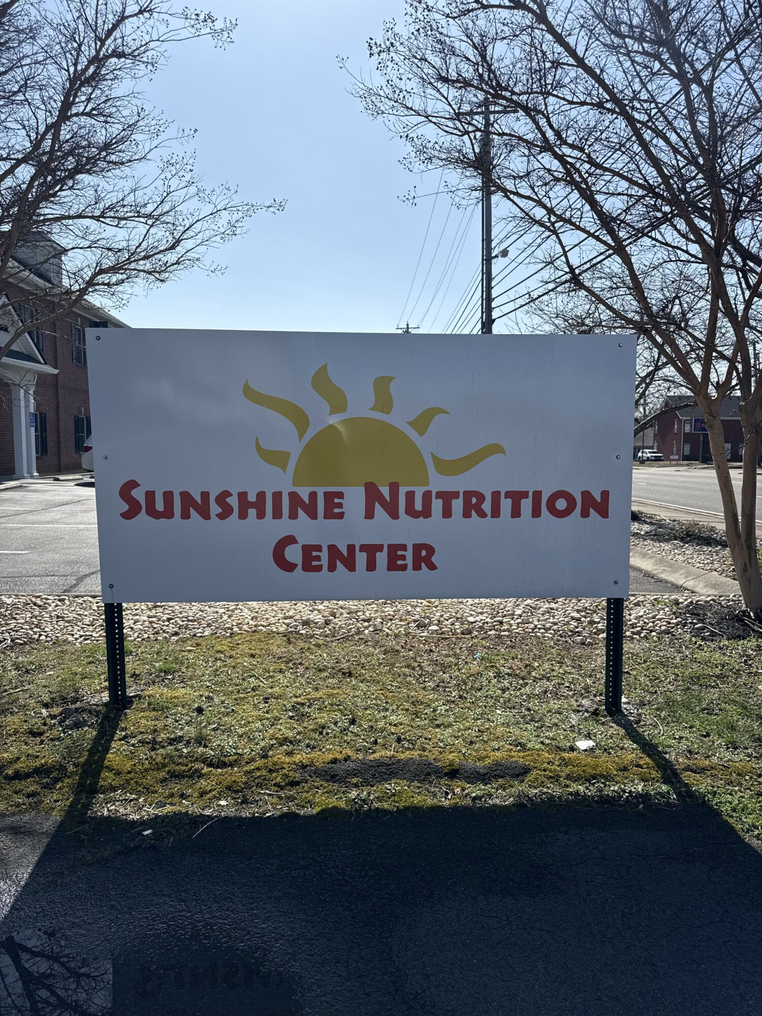 Sign for Sunshine Nutrition Center with a sun logo, outdoors on a sunny day, trees in the background.