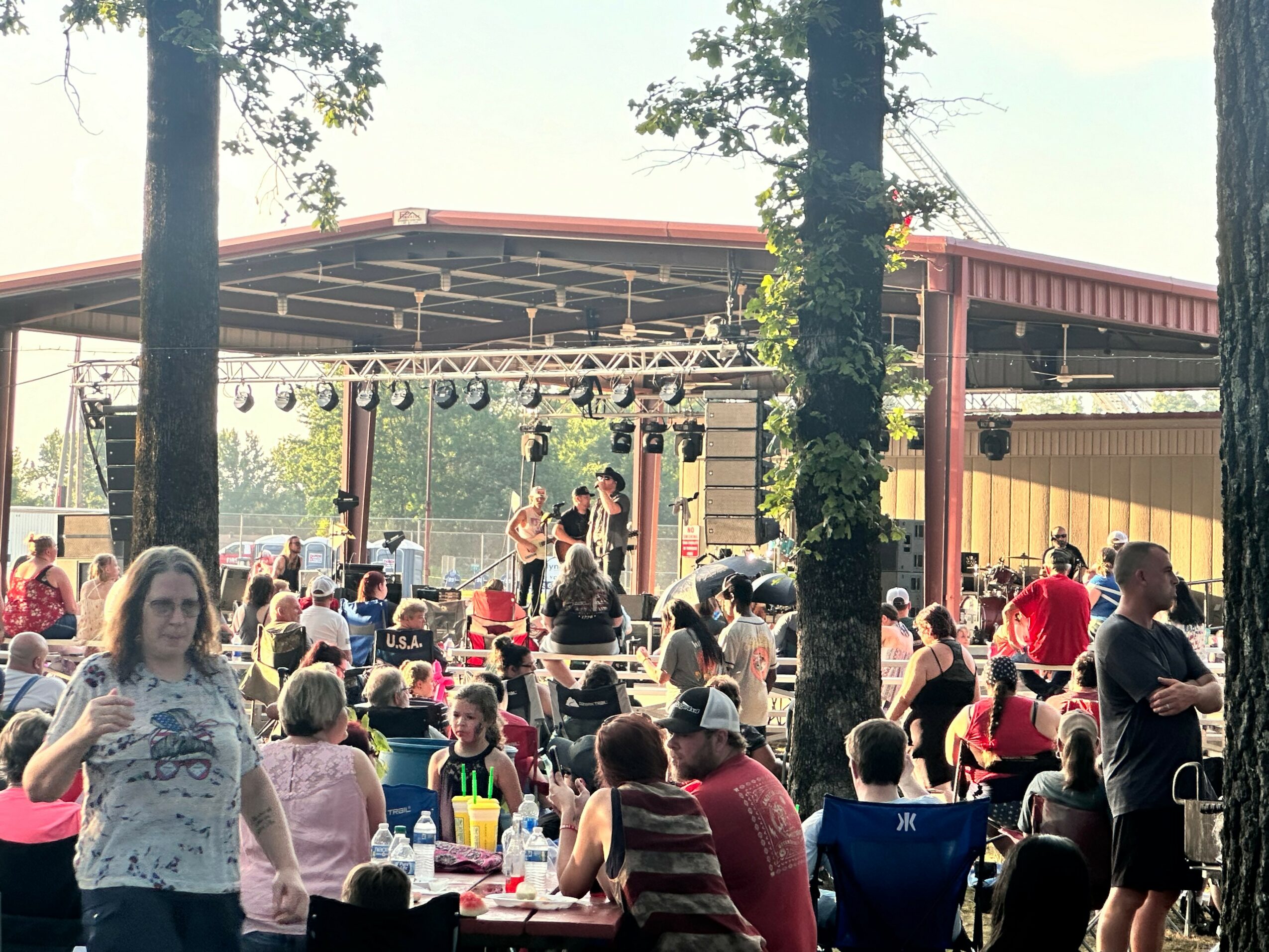 Outdoor event with a stage, audience seated at tables, trees, and a pavilion structure, daytime setting.