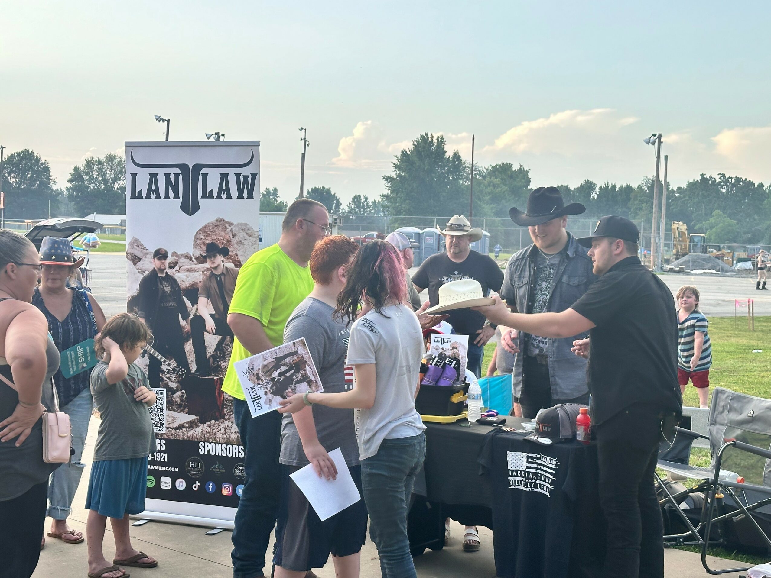 People gather outdoors near a table with signs and banners, some holding papers, with a parking lot and trees in the background.