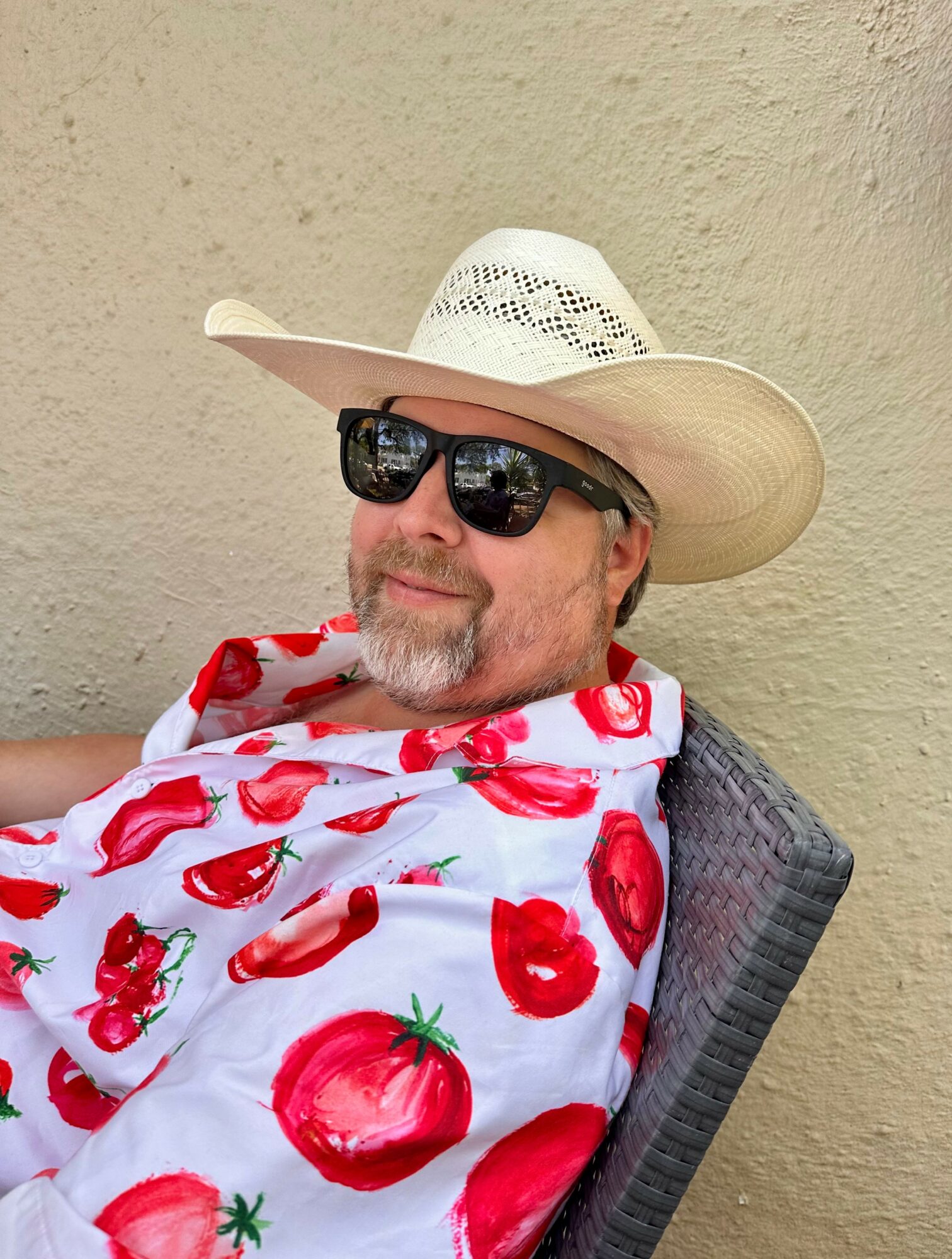 Man wearing sunglasses, a wide-brimmed hat, and a shirt with red tomatoes, sitting against a beige wall.