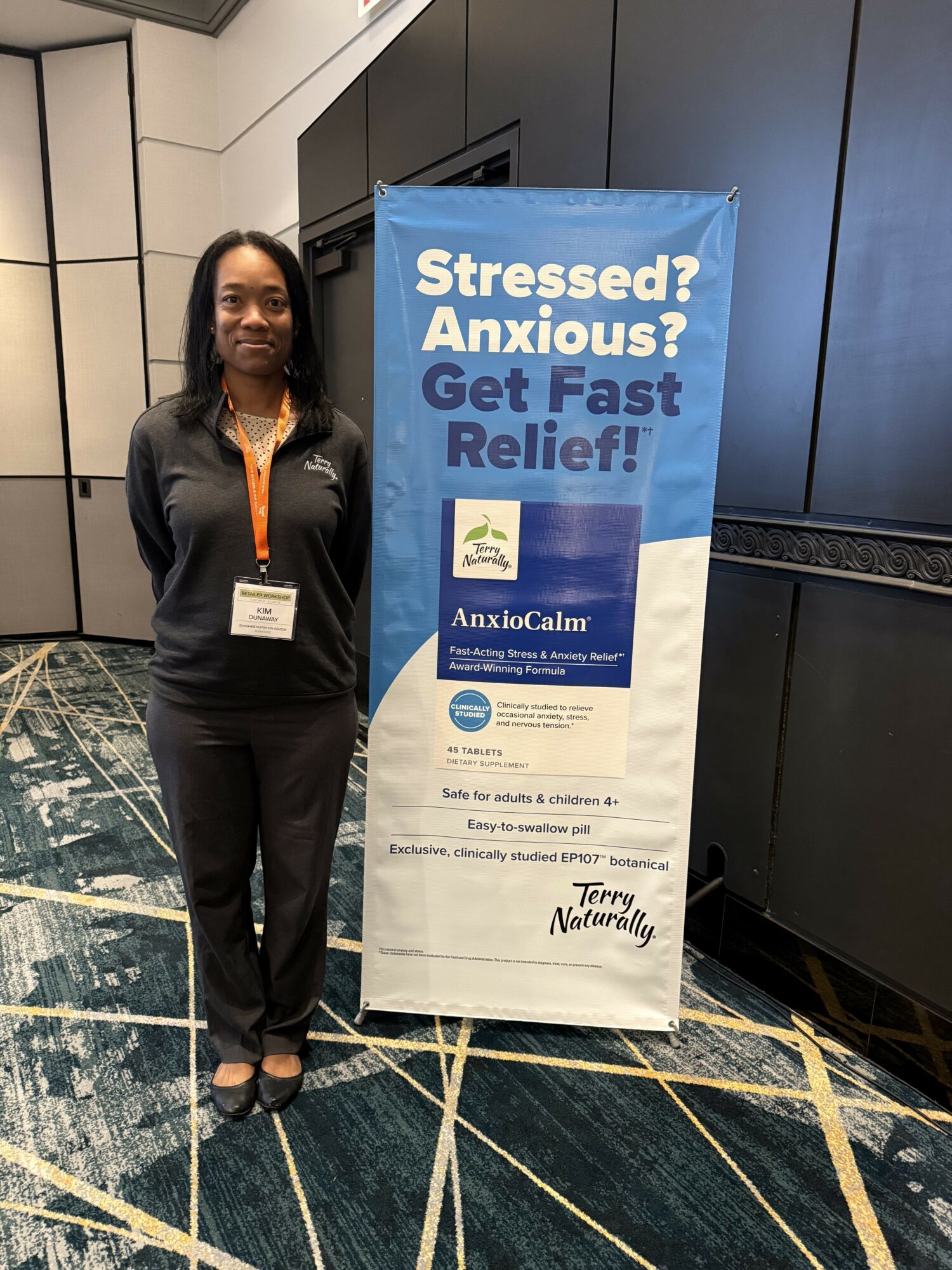 Woman standing next to a blue and white banner promoting stress and anxiety relief at a conference, in a modern indoor setting.