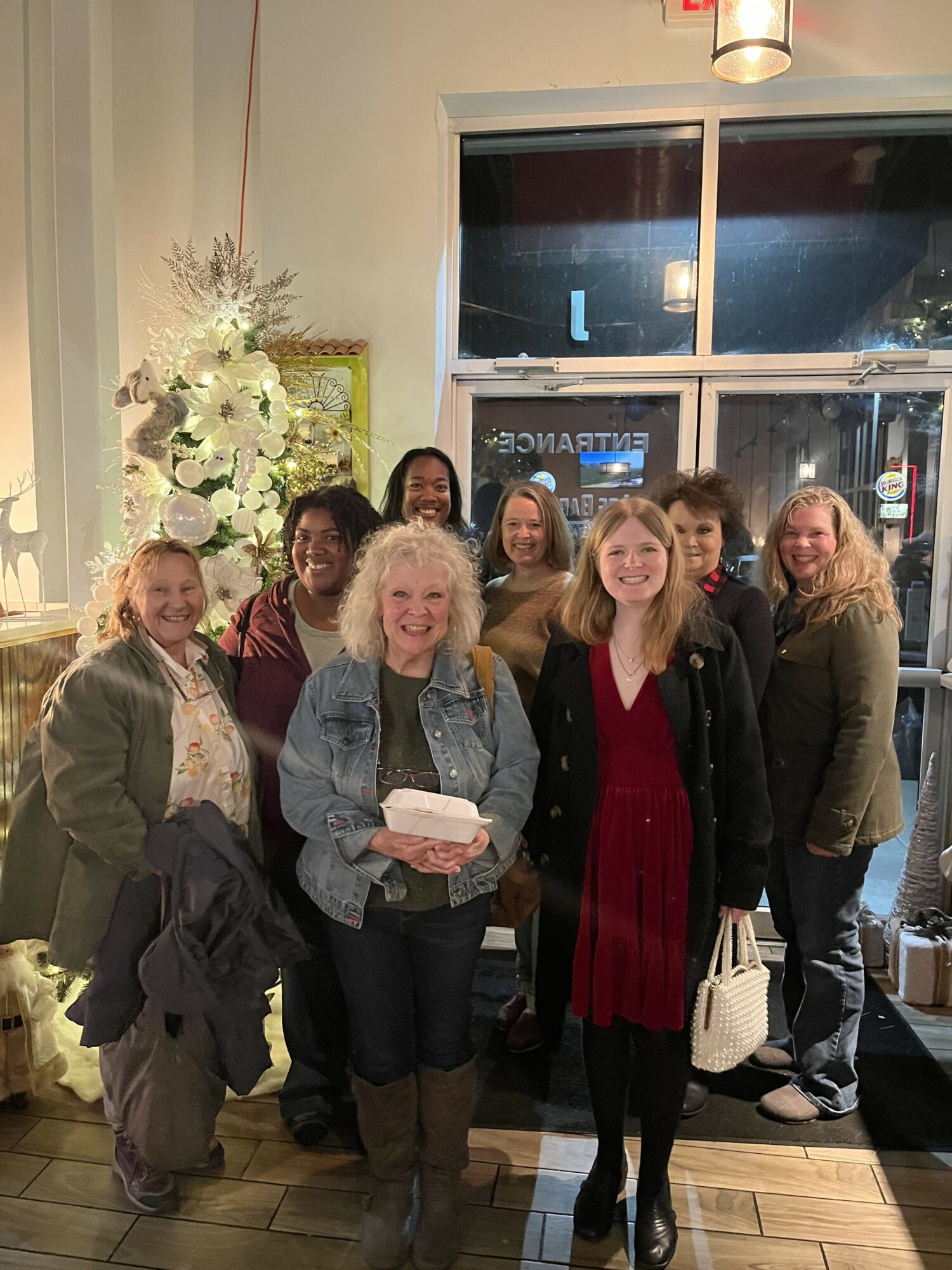 Group of nine women standing indoors near a large window with a flower arrangement, smiling for the photo.
