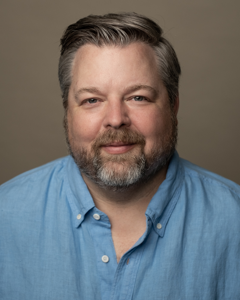 Portrait of a middle-aged man with gray hair and beard, wearing a blue button-up shirt, smiling softly, neutral background.