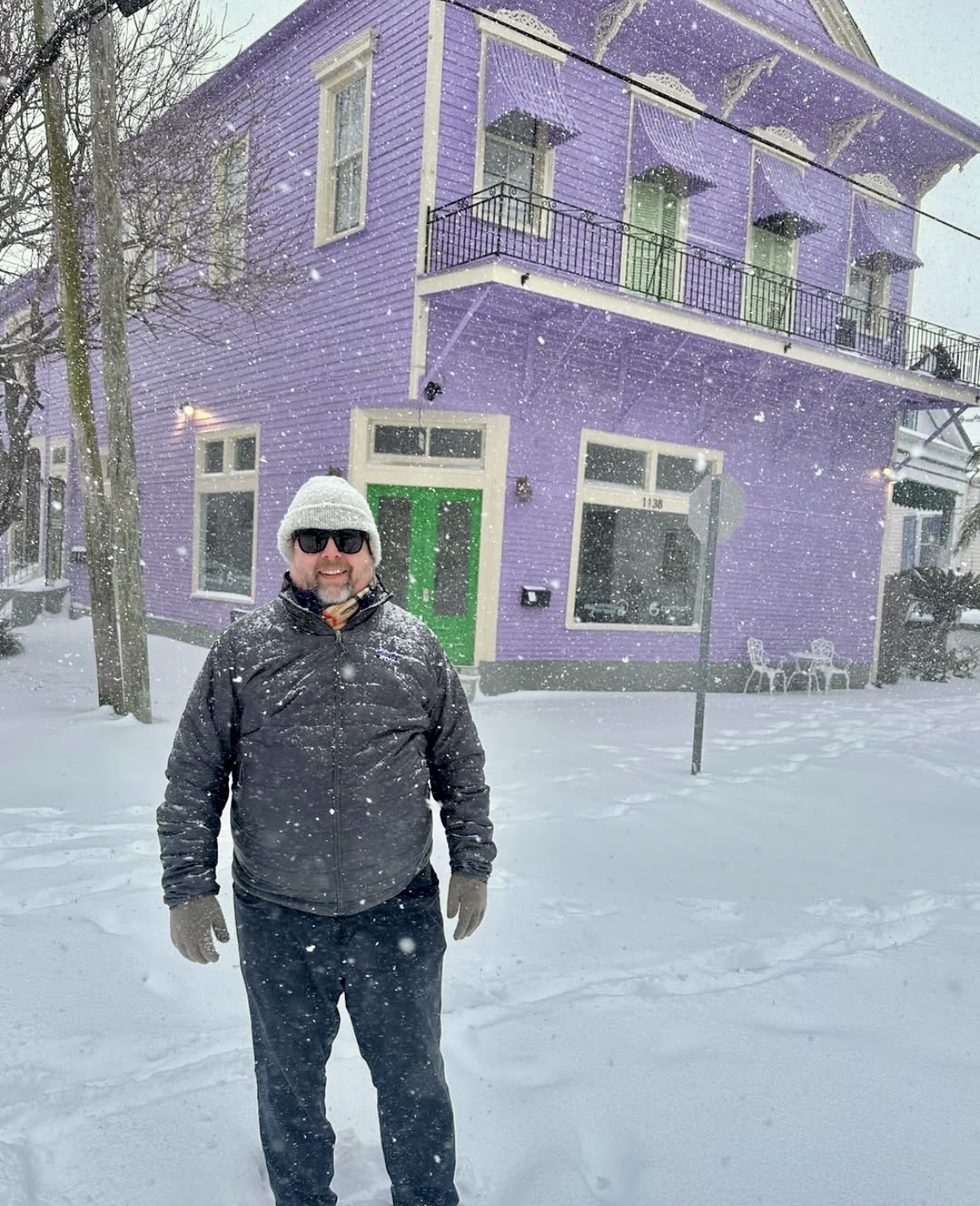 Person wearing sunglasses and winter clothing standing in snow in front of a purple building with green door and windows.