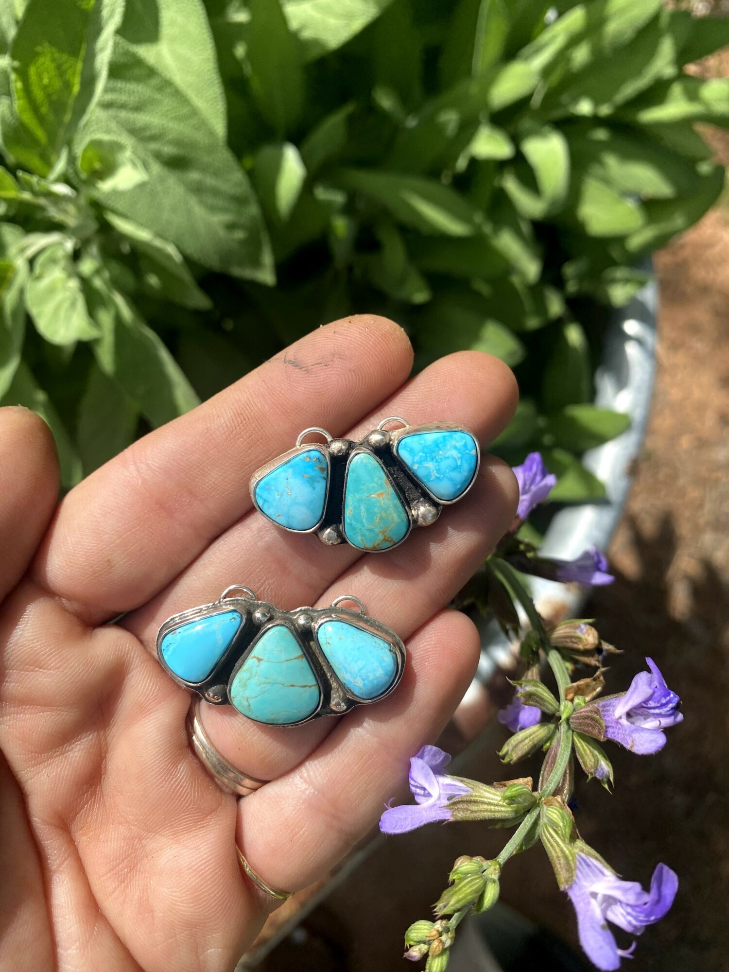 Hand holding two rings with turquoise stones, surrounded by green leaves and purple flowers, outdoors.