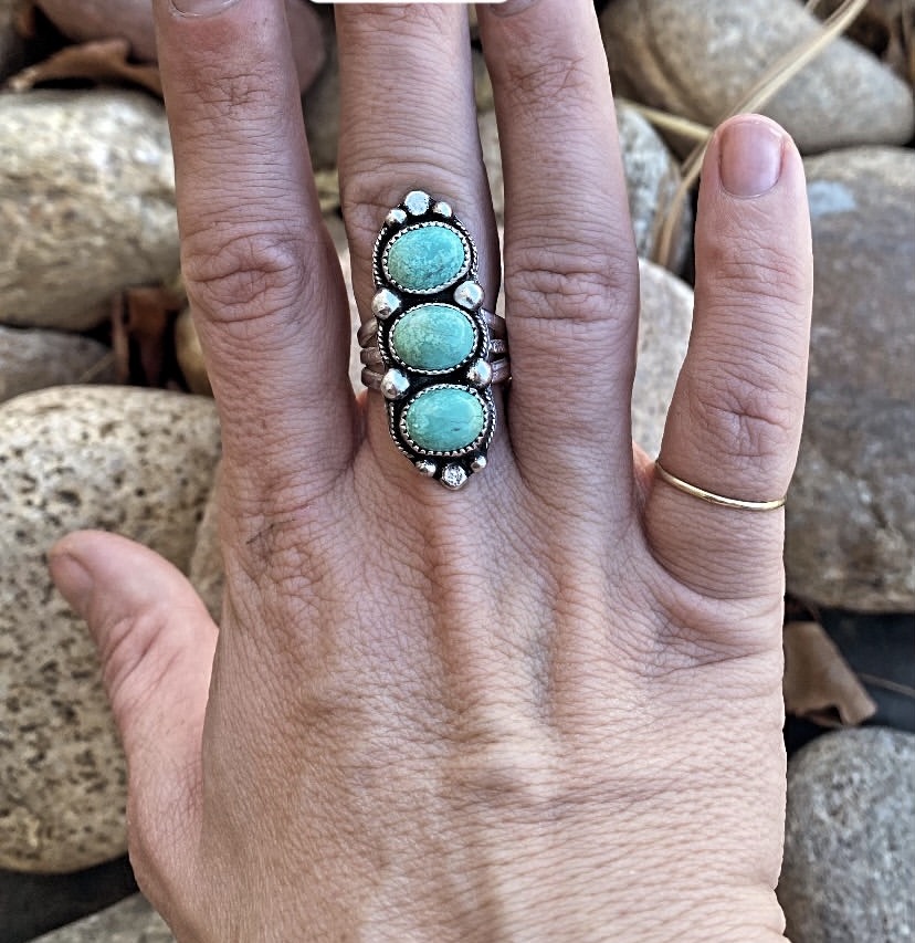 Hand with a silver ring featuring three turquoise stones, surrounded by small silver accents, against rocks background.
