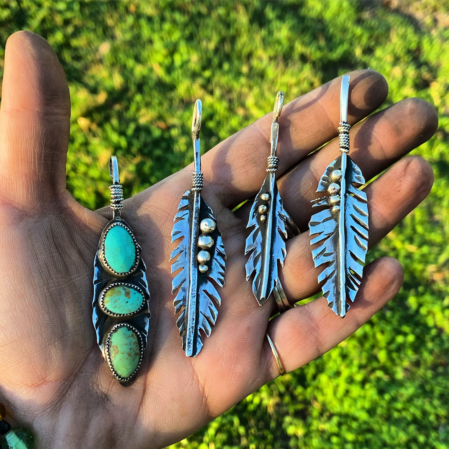 Hand holding four silver earrings with turquoise and feather designs, outdoors with green foliage background.