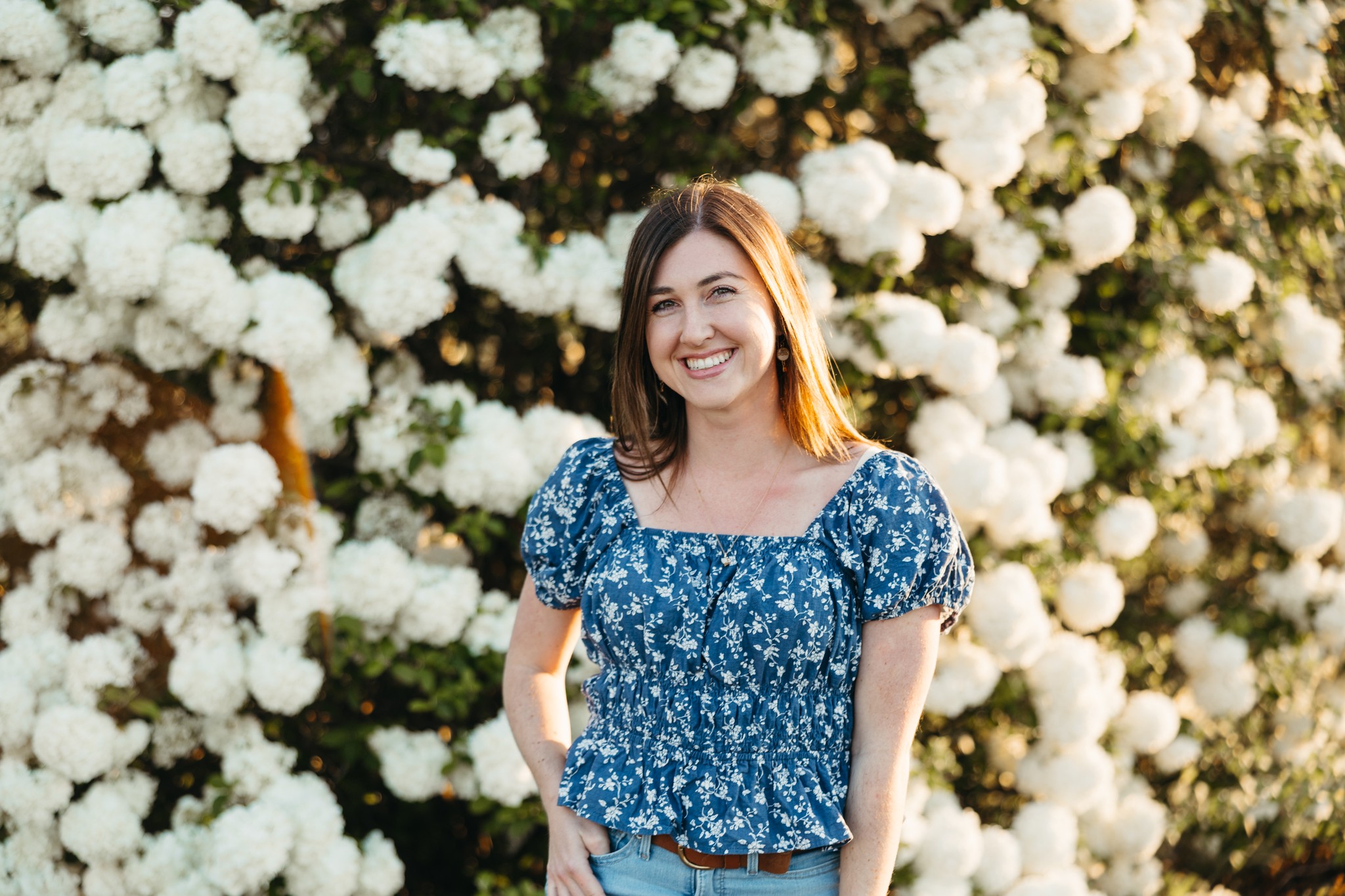 Smiling woman in a blue floral dress standing in front of a large white flower bush outdoors.