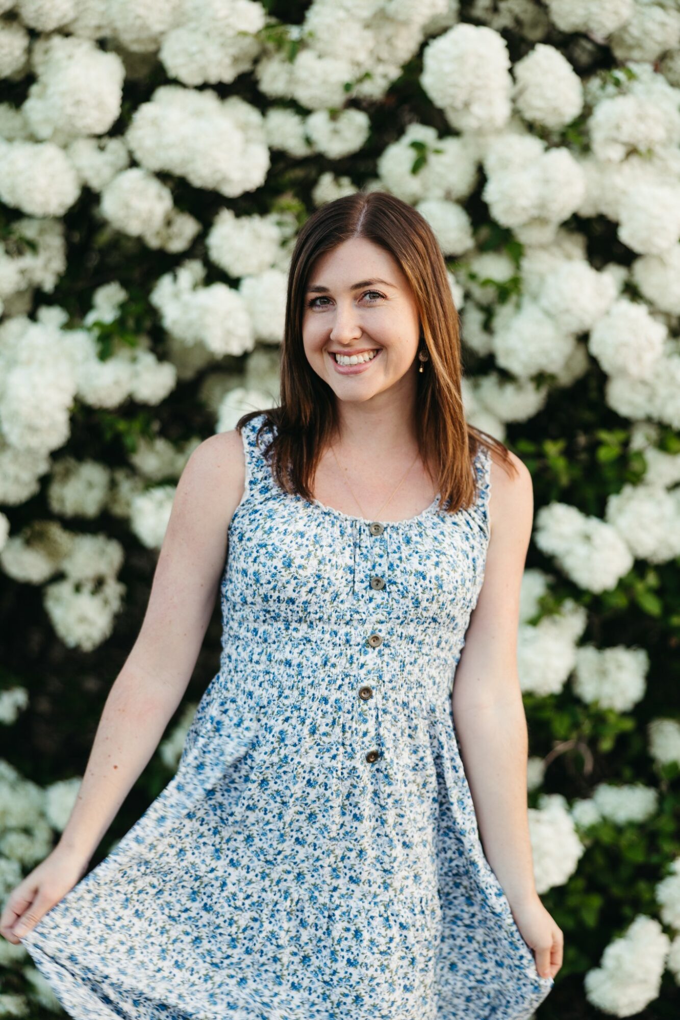 Young woman with brown hair smiling in front of white flowering bushes, wearing a blue and white patterned dress.
