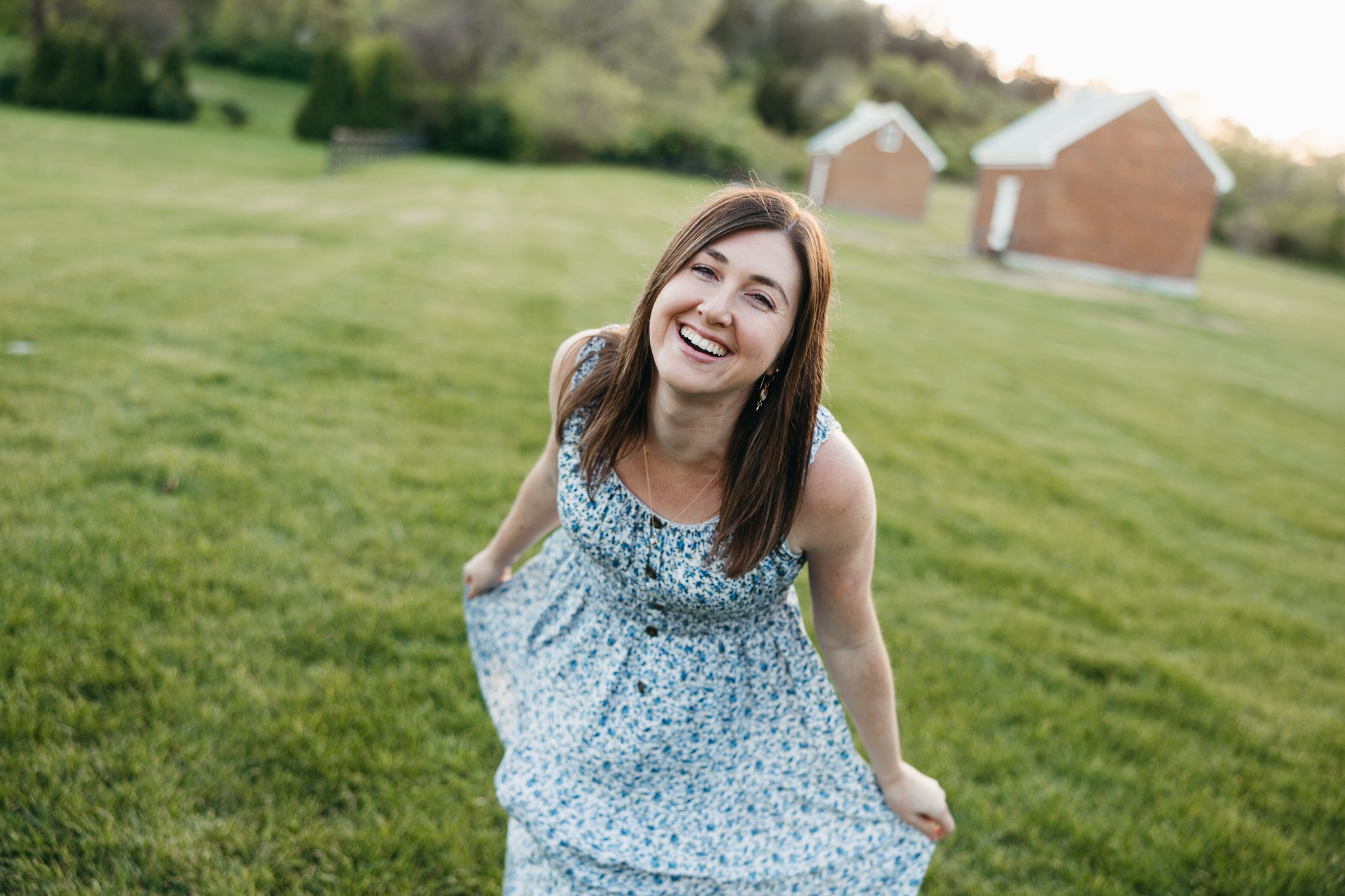 Young woman with long brown hair smiling outdoors in a grassy area, wearing a light blue dress with small patterns.